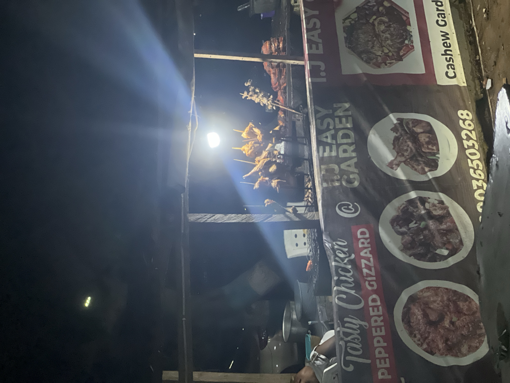 The image shows a food stall in Abuja, Nigeria, at night. Skewered meat, likely chicken, is being grilled over a light source, illuminating the scene. To the right of the grill is a sign for "I.J Easy Garden," advertising "Tasty Chicken" and "Peppered Gizzard." The sign also lists "Cashew Garden" and a phone number. A person is partially visible at the bottom of the image, near a metal pot or container. The environment appears to be a street or roadside setting.