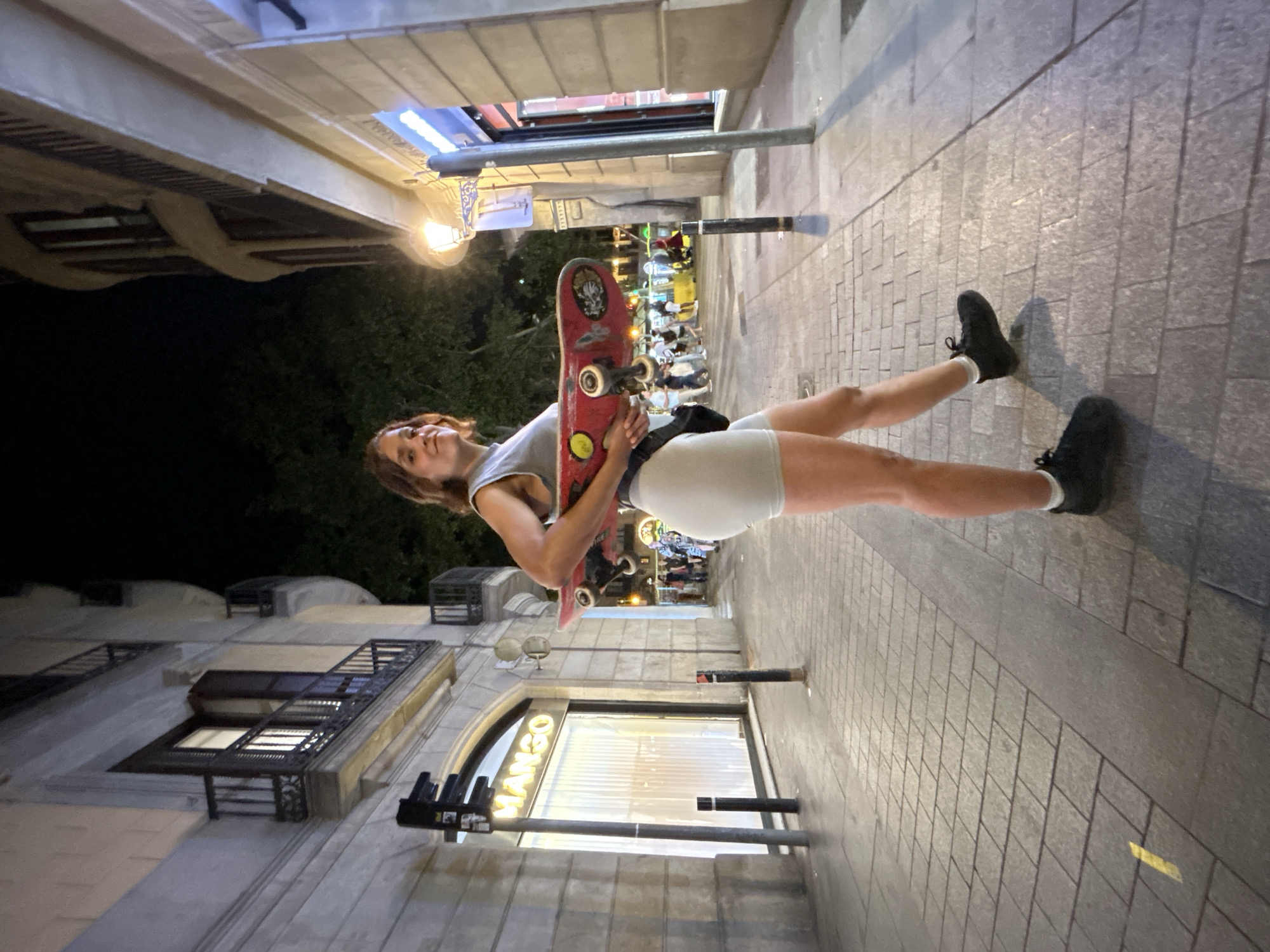 A young woman with short brown hair is holding a red skateboard under her left arm while standing on a cobblestone street at night. She is wearing a light gray tank top, light gray bike shorts, white socks, and black sneakers. A black fanny pack is visible around her waist. The woman is looking towards the camera with a slight smile.
Behind her, a building with a lit store window displaying the "MANGO" logo is visible. To the right of the woman, another building with balconies and windows is illuminated by streetlights. In the background, other pedestrians are walking along the street, and trees are visible. The scene appears to be a commercial street in a city.
This location is likely La Rambla in Barcelona, Spain, given the presence of the Mango store and the architectural style of the buildings.