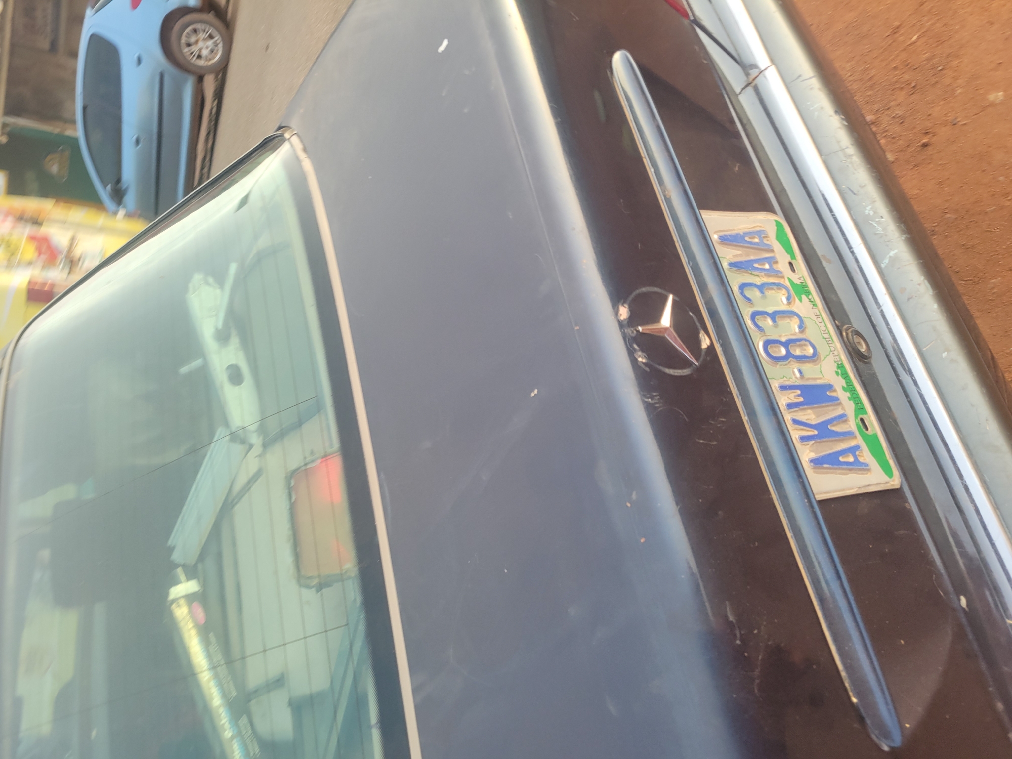 The image shows the rear of a dark-colored Mercedes-Benz. The license plate reads "AKW-833AA" and appears to be from the Federal Republic of Nigeria. The location is Unknown, Unknown. Other details include the car's rear window, the Mercedes-Benz logo, and a partial view of a light blue car in the background. The car appears to be in an urban environment.