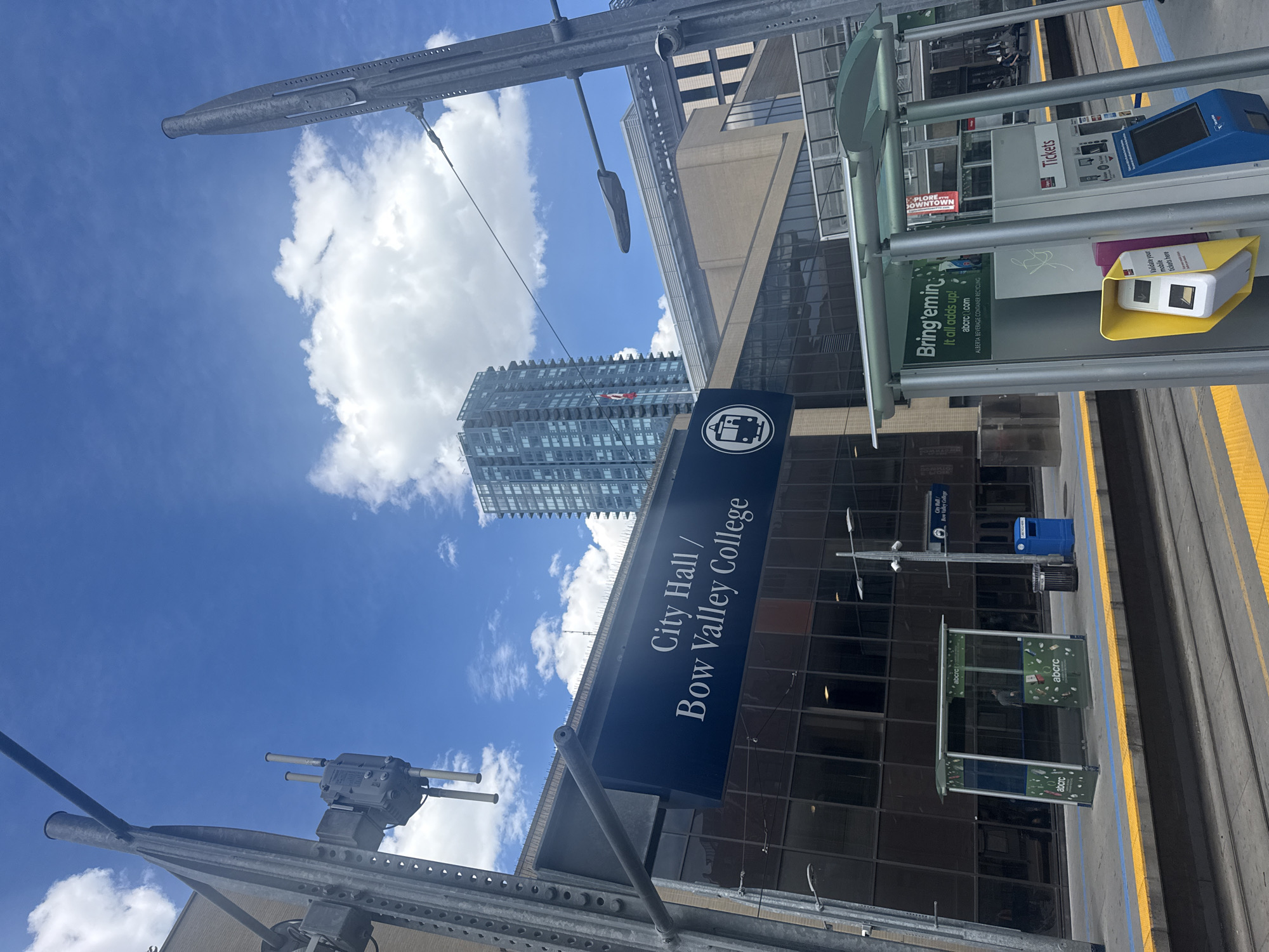 The image shows a Calgary C-Train station with a sign that reads "City Hall / Bow Valley College". The station platform has yellow and blue lines indicating safety boundaries. There are tracks for the C-Train and a small shelter with advertisements for "abcrcc.com". A ticket vending machine is visible on the platform. In the background, there is a modern, tall building with a blue facade, and other city buildings. The sky is blue with scattered white clouds. There are no people clearly visible in the image. The scene depicts an urban transit environment in Calgary, Canada.