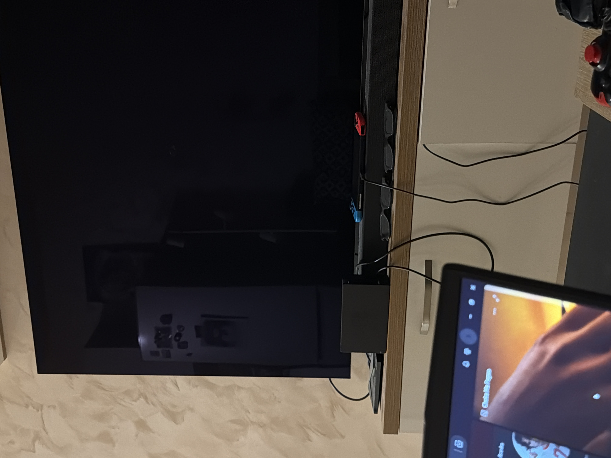 A close-up, eye-level shot shows a black television on a wooden cabinet. On the cabinet, to the left of the television, is a black gaming console and several cables. To the right of the console are two red and blue gaming controllers and two pairs of 3D glasses. A laptop is in the foreground, partially obscuring the cabinet and television. The wall behind the television has a textured, light beige finish. The image appears to be taken indoors in a living room.