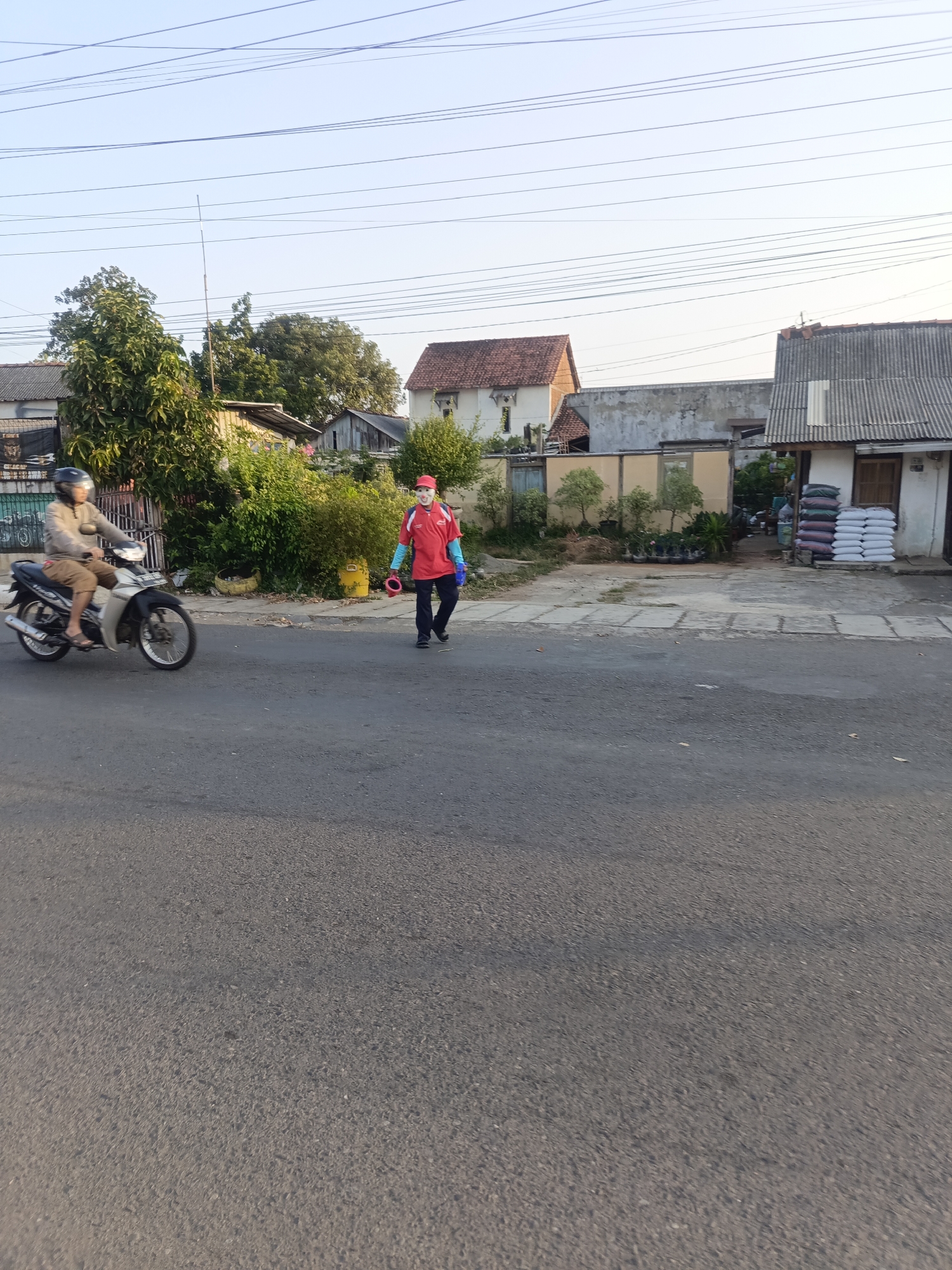 A man on a motorcycle is traveling down a paved road. He is wearing a light-colored jacket, tan shorts, and a black helmet. The motorcycle is silver and black. To the right of the road, a man in a red shirt and blue sleeves is walking. He is wearing a red hat and carrying two red objects. He appears to be wearing gloves. Behind him are houses, trees, and stacks of white and dark-colored sacks. The sky is clear and blue. The scene takes place in Desa Gunungsimping, Indonesia.