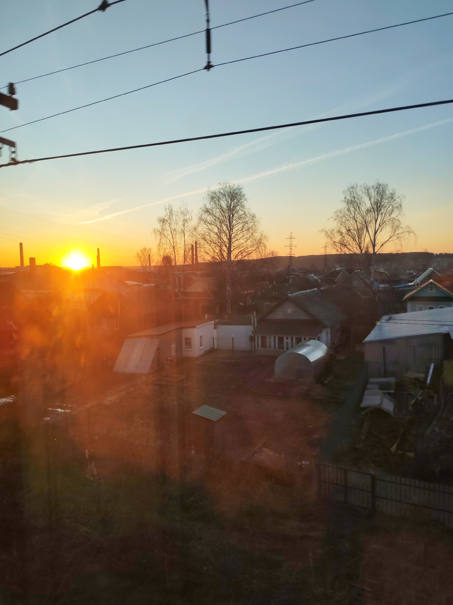 The image captures a vibrant sunset or sunrise over a semi-rural landscape, likely viewed from a moving vehicle due to the prominent overhead power lines and a subtle sense of motion. The sun, low on the horizon to the left, emits an intense golden-orange light that bathes the scene, creating a strong lens flare effect and reflections, particularly in the lower left foreground.
The setting features an assortment of residential buildings, including older wooden houses and more modern structures, interspersed with smaller outbuildings like sheds and tunnel-shaped greenhouses. Bare deciduous trees, indicative of late autumn or early spring, are scattered throughout, their skeletal branches silhouetted against the bright sky. In the background, on the left, industrial chimneys or smokestacks rise, suggesting a nearby industrial area. On the right, further back, several electricity pylons are visible. The sky transitions from a pale blue at the top to warm oranges and yellows near the horizon, with faint contrails streaking across it. The ground is largely bare earth or sparse, dry vegetation.
No people are visible, and no specific activity or event is taking place beyond the natural progression of daybreak or dusk. The strong, warm light is the most notable detail, providing a dramatic contrast with the muted tones of the landscape and structures. No visible text is present in the image.