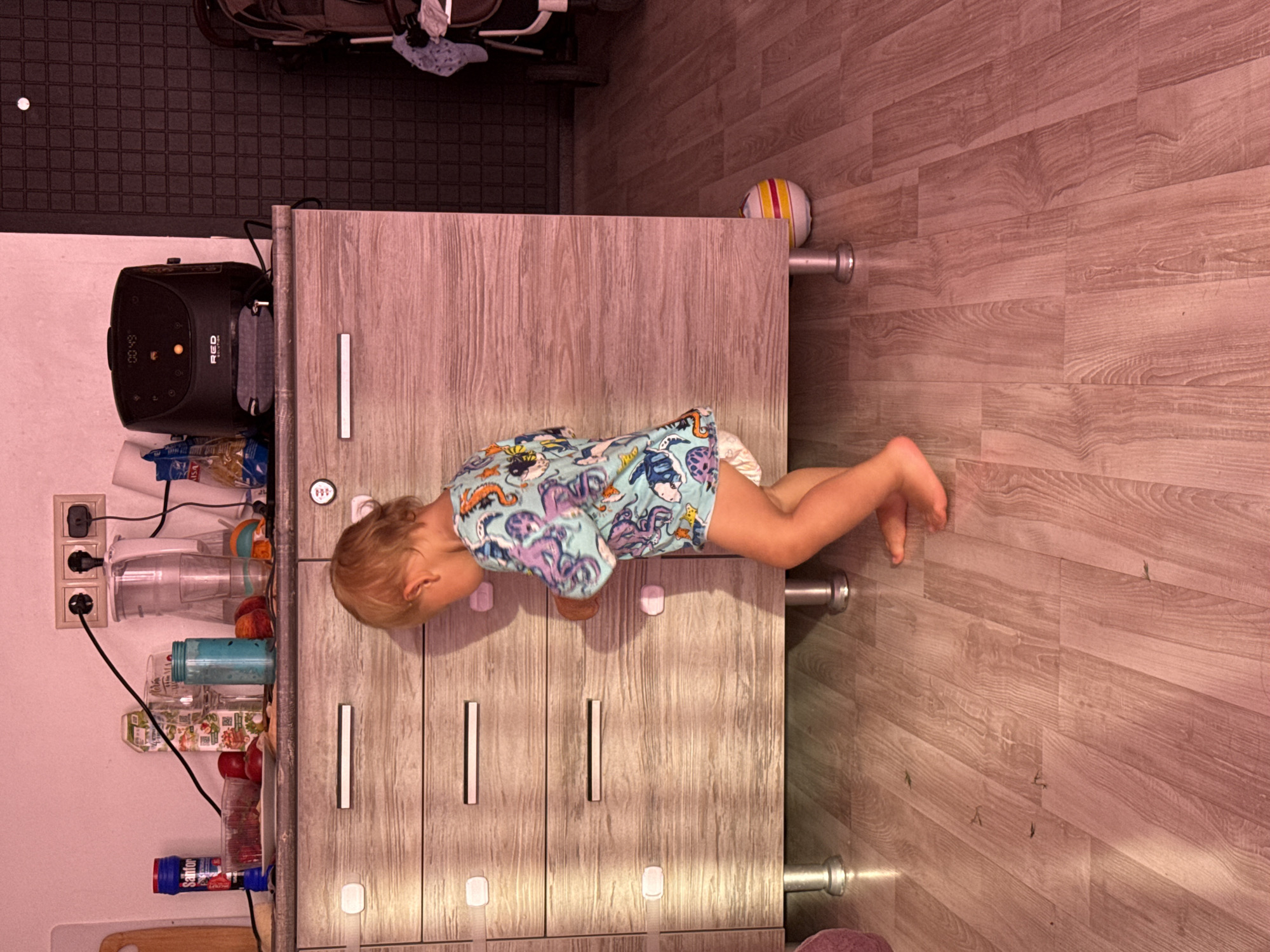 An indoor scene depicts a young child, possibly a toddler, wearing a light blue shirt with an octopus print and a diaper. The child is facing a wooden dresser with several drawers, some of which have child safety latches. The dresser is situated in what appears to be a kitchen or living area. On top of the dresser, various items are visible, including a black appliance with a digital display, a blender, and containers of food. The floor is made of wooden planks. A baby stroller is partially visible in the background. The lighting in the room is warm and somewhat dim, casting pinkish tones. The image does not contain any specific landmarks or indicators to suggest a location in the city of Sochi, Russia.