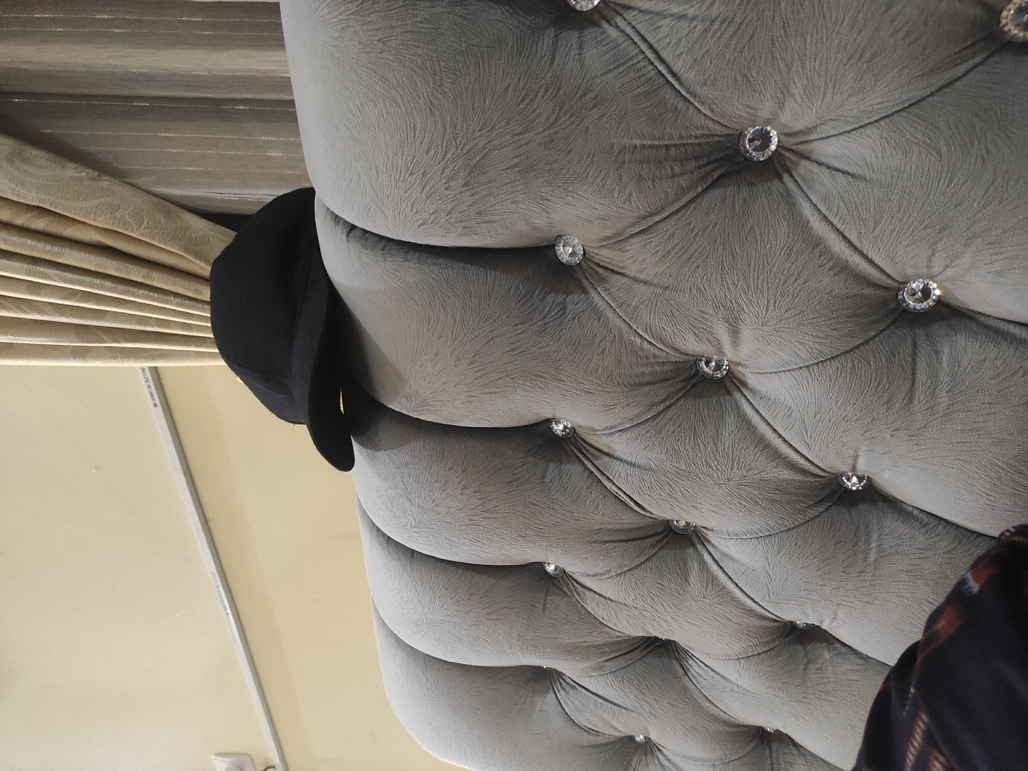 A black baseball cap is resting on the tufted headboard of a bed. The headboard is upholstered in a gray fabric with a subtle embossed pattern and decorated with clear crystal-like buttons. Behind the headboard, beige curtains are drawn. A fluorescent light fixture is mounted on the wall above the headboard, which is painted a pale yellow. The image was taken in Sumedang, Indonesia, within a bedroom environment. No people are visible in the frame.