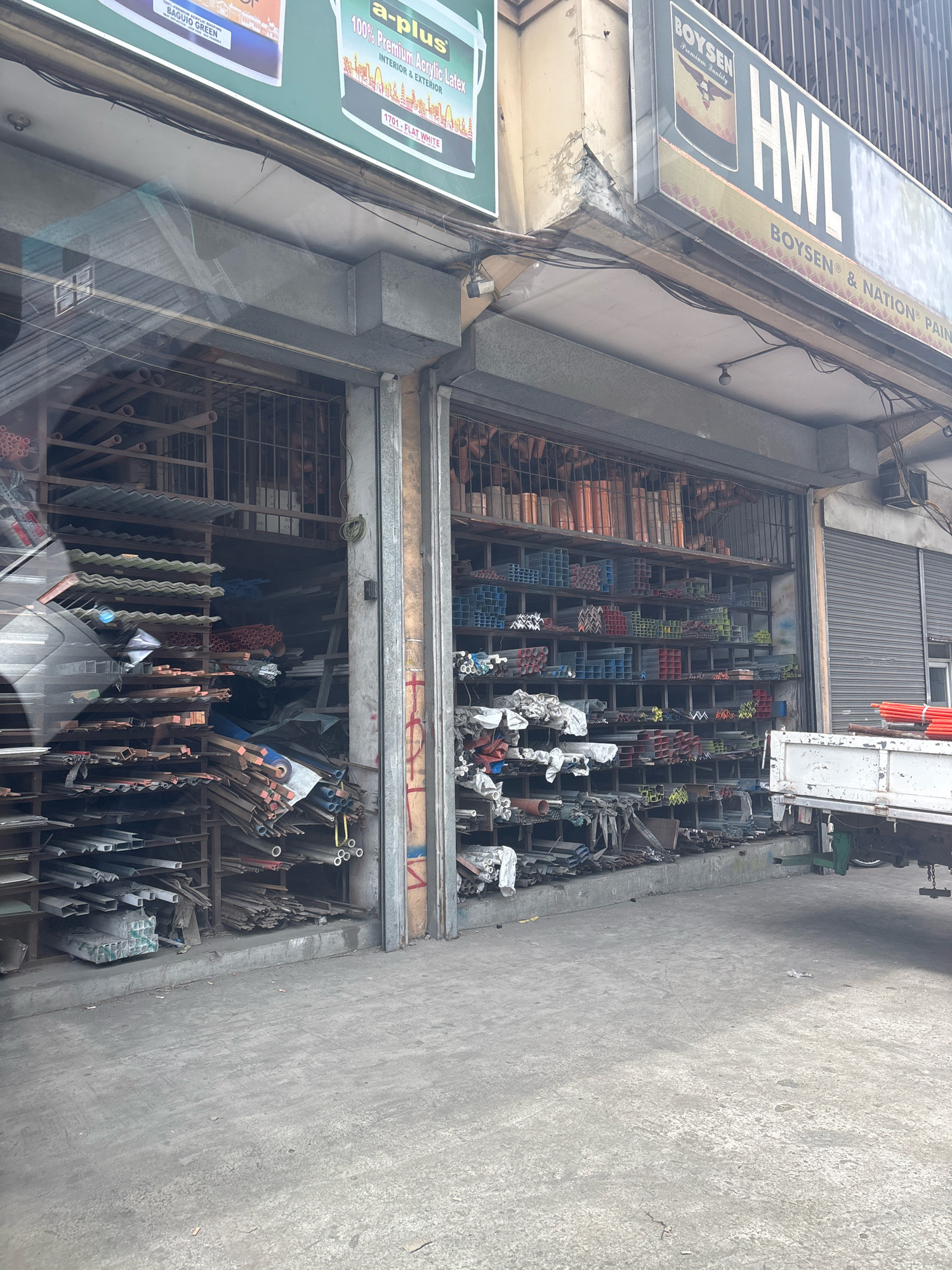 The image shows a hardware store in Carmona, Philippines, with its entrance open, revealing shelves stocked with various metal and construction materials. The store appears to be a wholesale supplier, indicated by the large quantities of items like metal pipes, rods, and sheets organized on shelves. A white truck is parked in front of the store, possibly for loading or unloading goods. The building facade has signs that are partially visible, including "HWL" and "BOYSEN," suggesting it is part of a chain or deals with specific brands. The street in front of the store is paved and appears to be an urban setting, with some debris on the ground. The overall impression is of a busy commercial establishment in a Filipino city.