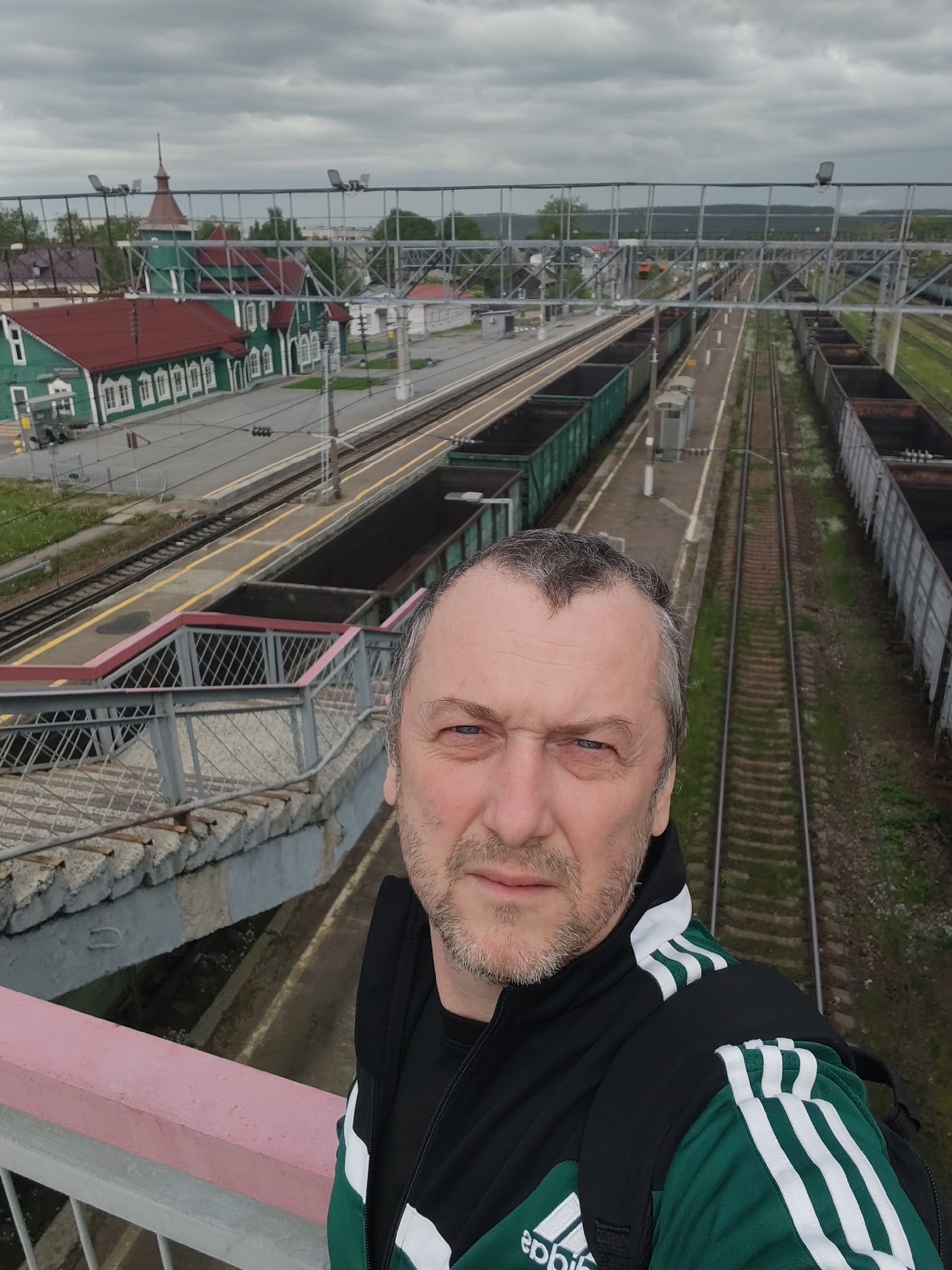 The image features a selfie of a middle-aged man positioned on a pedestrian bridge, overlooking a railway station in Medvezhyegorsk, Russia. The man, with short, greying hair and a light beard, faces the camera with a neutral to slightly serious expression. He is wearing a black and green Adidas tracksuit top with prominent white stripes on the sleeves and shoulders, and a black t-shirt. A black backpack is also visible on his shoulders.
The scene behind him is a railway yard and station under a cloudy, overcast sky, suggesting daytime. Below the bridge, multiple railway tracks stretch into the distance. To the left, a charming, distinctively Russian wooden railway station building painted green with a red roof and a small, pointed spire stands out. Its white-framed windows provide a strong contrast against the green.
Alongside the platforms, several freight train wagons are visible; some are green, open-top wagons, while others appear darker and potentially loaded with bulk materials like coal or ore. Overhead electrical lines and their supporting gantries crisscross the railway area. In the background, beyond the tracks and buildings, there are trees and distant hills, indicating a semi-rural or natural setting surrounding the town. The overall impression is that of a functional railway hub on a typical overcast day. The only visible text is the "adidas" logo on the man's jacket.