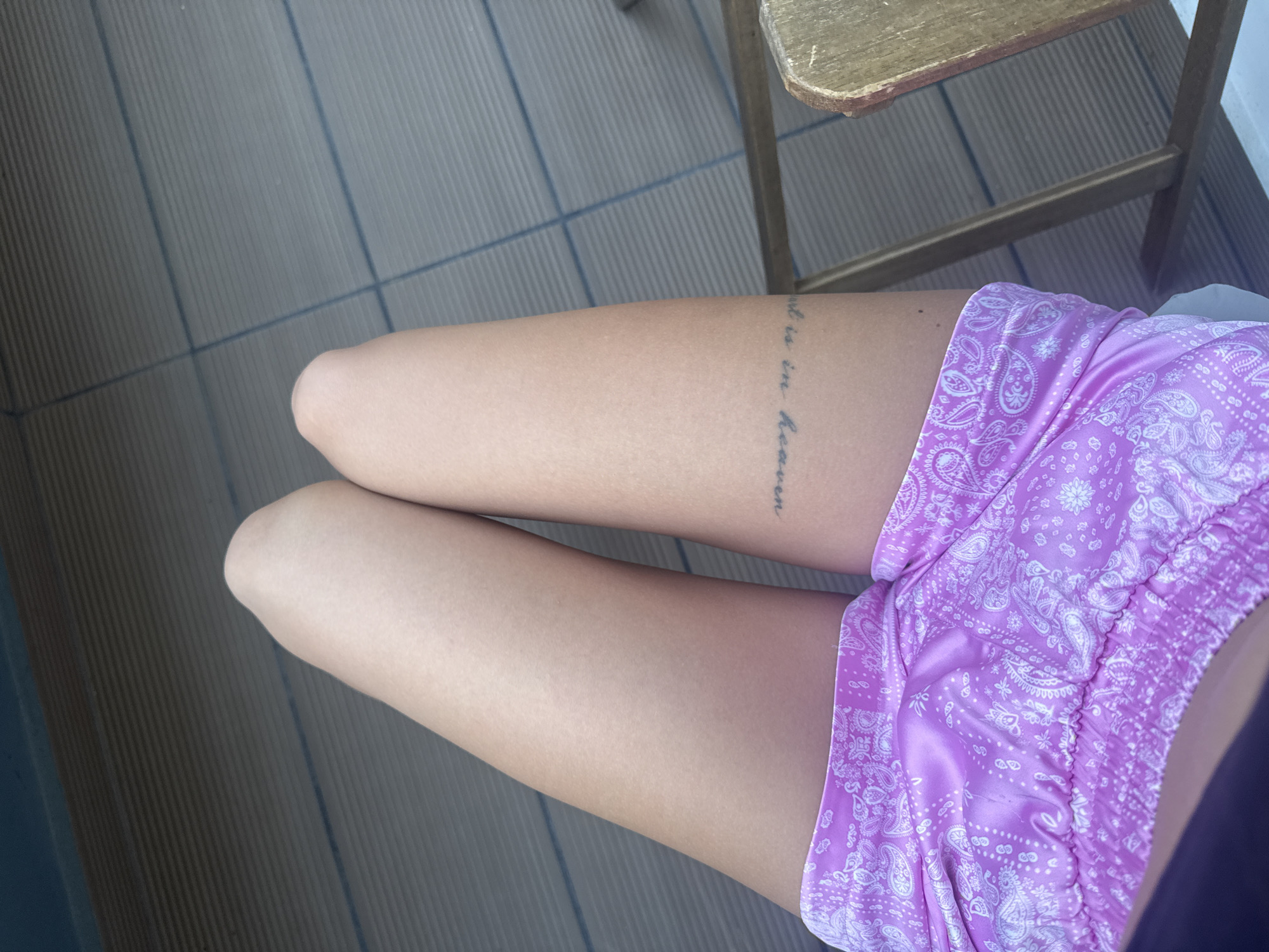 The image captures a close-up, high-angle view of a person's legs, from the thighs to just above the knees, as they are seated. The person is wearing bright purple shorts with a white paisley pattern. A tattoo is visible on the outer side of the left thigh, written in vertical script, part of which reads "...is in Heaven". In the background, there is a patterned floor, likely made of narrow planks or tiles, and the lower portion and a small part of the wooden seat of a rustic wooden chair are visible in the upper right. The lighting is bright, suggesting it is daytime. The setting appears to be outdoors, possibly on a balcony or patio, in Plovdiv, Bulgaria.