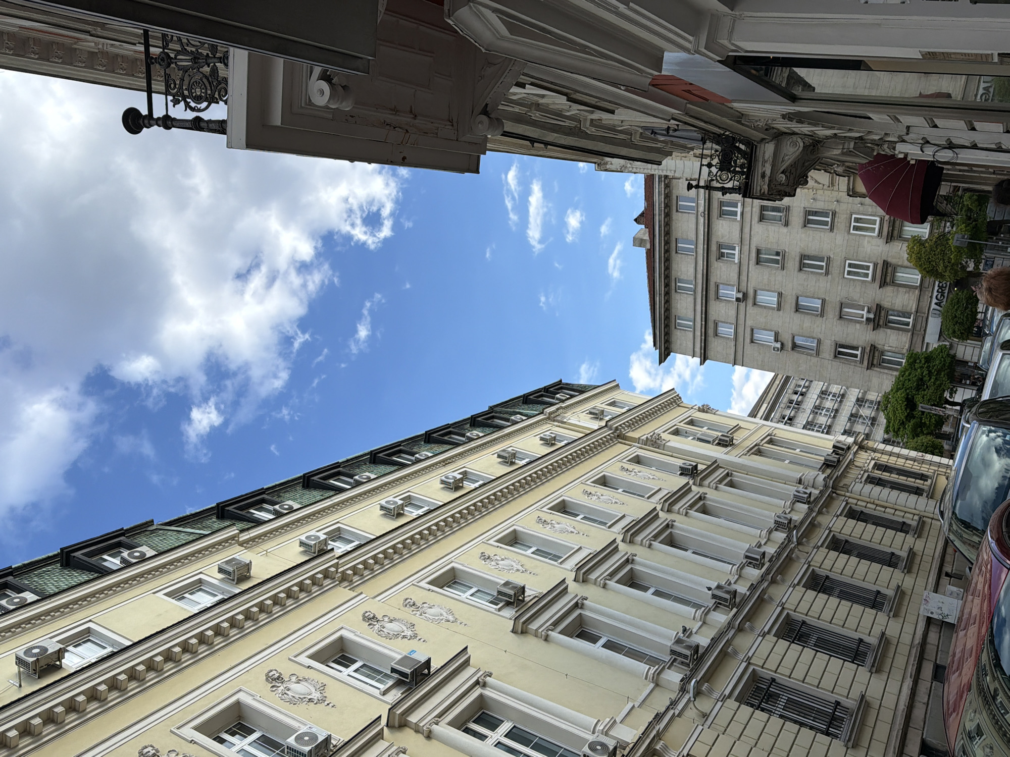 The image, taken from a low angle looking upwards, captures a view between two rows of multi-story buildings on an urban street during bright daylight.
On the left, a tall, light yellow building dominates the frame, featuring elaborate classical architectural details such as ornate window frames, decorative relief carvings between floors, and prominent cornices. Numerous external air conditioning units are visible below and above many of its windows. The building is topped with a distinctive dark green tiled mansard roof.
On the right, a taller, less ornate grey building stands, characterized by a uniform grid of rectangular windows. A red retractable awning extends from one of its lower levels. A sign on this building displays the partial text "AGREB". In the background, between the two main structures, another building is partially visible.
The sky above is a vibrant blue with scattered, fluffy white cumulus clouds, indicating fair weather. The street level, partially visible at the bottom, shows reflections on parked cars. A small, illegible white sign is affixed to the wall of the yellow building near the bottom. No specific activity is taking place, and only a hint of a person's head is visible near the grey building.