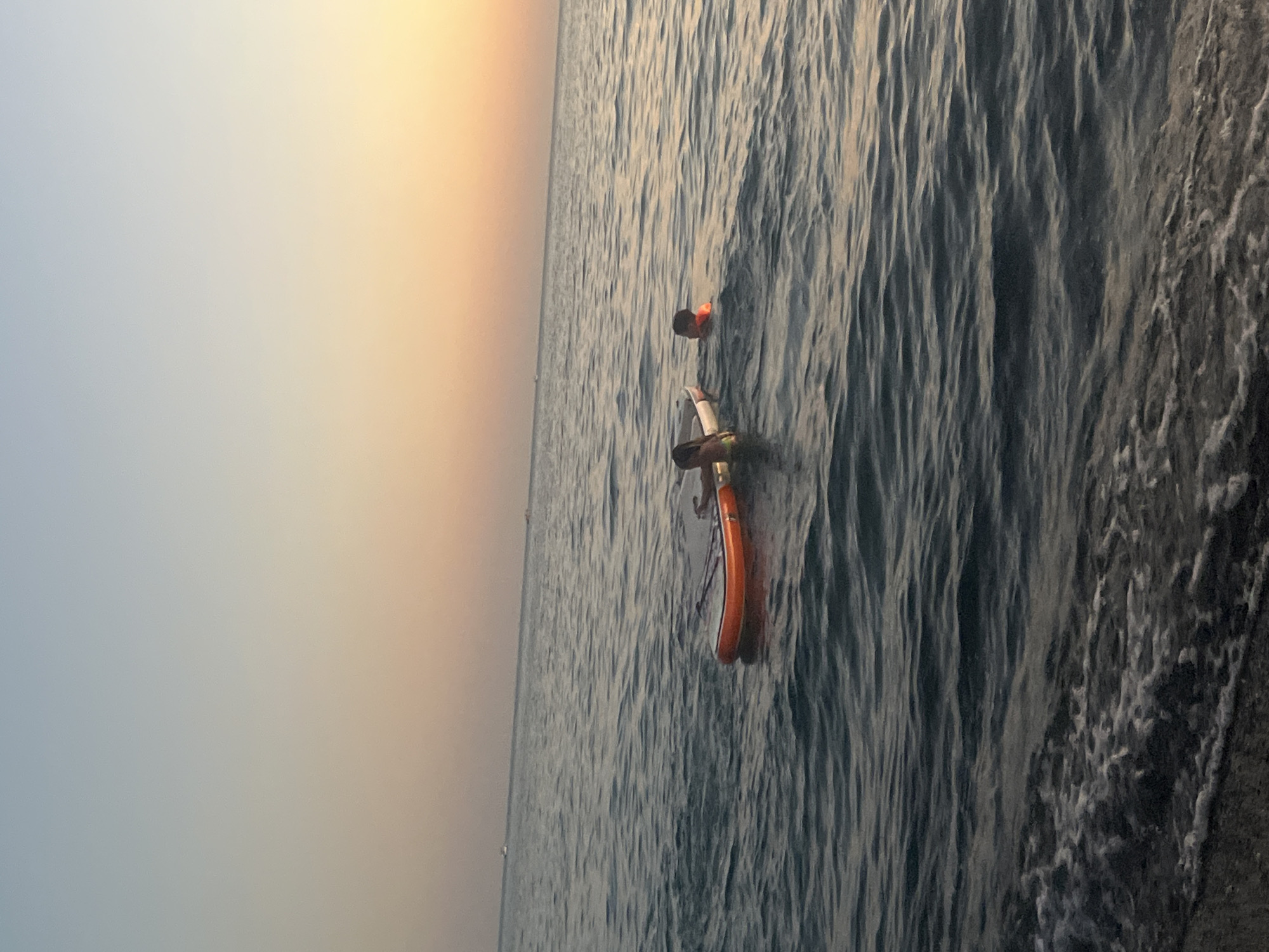 Two children are in the sea near an inflatable raft at sunset. One child is on the raft, and the other is in the water beside it. The sky is a gradient of pale blue to orange, indicating a sunset. The sea is calm with gentle waves. In the distance, several boats are visible on the horizon. The foreground shows the edge of a beach with small waves lapping the shore. The scene appears to be a serene evening at a beach in Sochi, Russia.