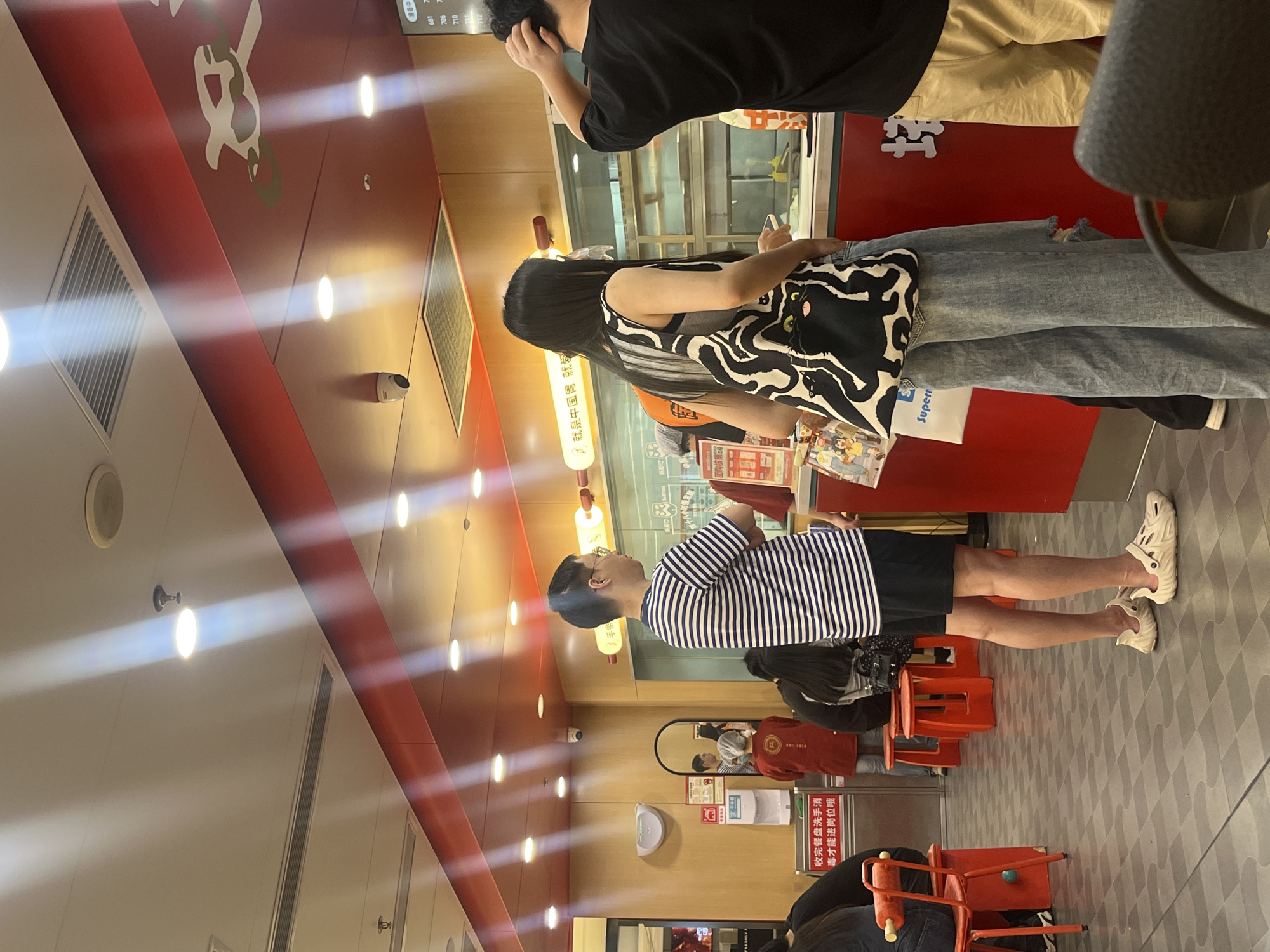 The image captures the interior of a casual Chinese fast-food establishment, reportedly located in Yuzhong District, China. The floor features a pattern of alternating dark gray and light gray square tiles. The lower walls are light wood paneling, transitioning to a prominent red counter area and a red ceiling section above it. The ceiling is equipped with multiple recessed spotlights, casting distinct light rays, along with several air conditioning vents and white dome-shaped surveillance cameras. A large, stylized white graphic is visible on the red ceiling section.
In the mid-ground, three individuals are standing near a red service counter. A man on the left wears a horizontal blue and white striped t-shirt, black shorts, and cream-colored, textured slip-on clogs. He stands with his arms crossed, facing right. Adjacent to him, a woman with long dark hair, wearing a black and white abstract patterned sleeveless top, light-wash wide-leg jeans with rips, and a clear plastic face shield, stands facing the counter while holding a mobile phone. She carries a black and white tote bag featuring a black cat design with green eyes. To her right, a man in a black t-shirt and tan shorts is partially visible, with his right hand resting on the back of his head.
In the background, near a wooden wall, a person in a red uniform and a white hairnet is visible, reflected in a large mirror. A wall-mounted hand sanitizer dispenser is next to the mirror, and a sign with Chinese text "收餐盘洗手消毒才能进岗位哦" (Wash hands and disinfect the tray before entering the station oh) is displayed below it. Several red stools and chairs with red frames are arranged in the seating area to the left. Behind the main counter, a digital display shows numbers such as 681, 706, 710, and 712, likely indicating order status. A sign above the counter reads "就是中国菜" (It's Chinese food).