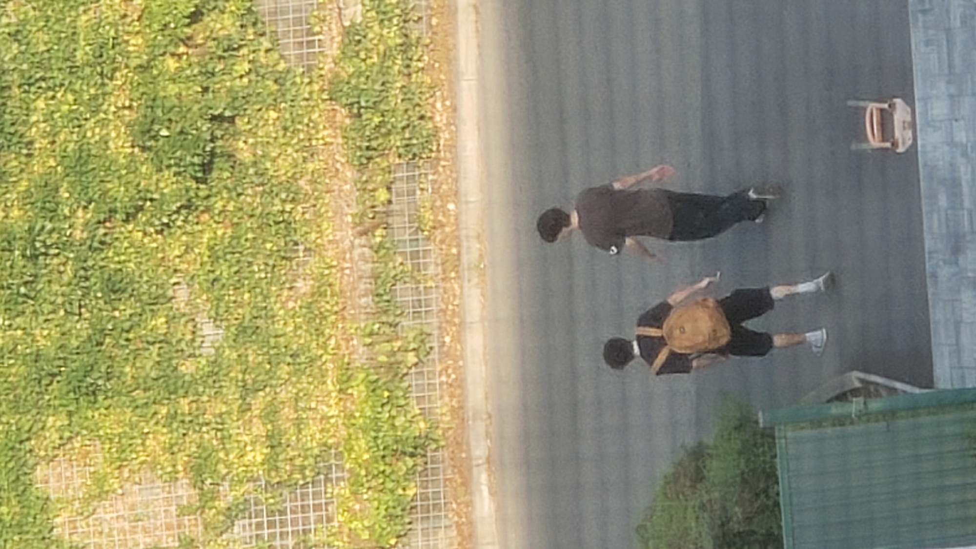Two individuals are walking away from the camera on a paved surface. The individual on the left is wearing a dark t-shirt, black shorts, white socks, and light-colored sneakers. They are carrying a tan backpack. The individual on the right is wearing a dark t-shirt and dark pants, with light-colored shoes. Behind them, a metal fence covered with green and yellow foliage runs along a slightly elevated embankment. In the foreground, a green structure is visible on the left, and a light-colored chair is on the right. The scene appears to be an outdoor urban environment, possibly in Prešov, Slovakia, given the context.