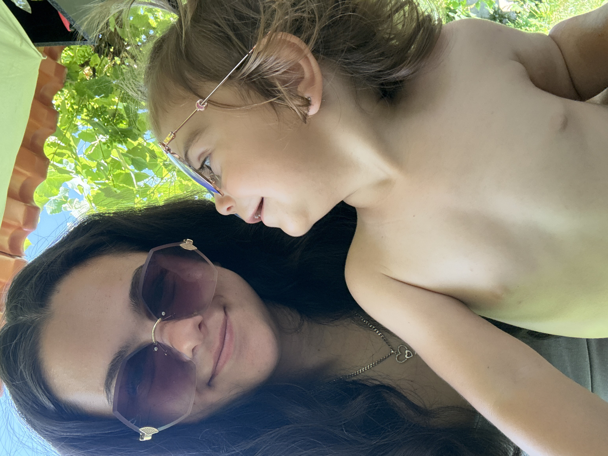 A close-up, selfie-style image features two people in an outdoor setting. In the foreground, a woman with long, dark hair wears large, hexagonal sunglasses with a rose-gold frame. A delicate silver necklace with a heart-shaped pendant is visible around her neck. Her gaze is directed upwards and to the right, with a slight smile on her face.
Positioned behind and slightly above the woman is a young child, also looking upwards and to the right. The child wears small, rose-gold framed glasses with a subtle heart detail on the side. The child's back is to the camera, revealing their bare shoulder and upper back.
The background consists of lush green foliage and bright blue sky, suggesting a sunny day. The lighting indicates it is daytime, likely late morning or afternoon. Hints of a light green object and a red object are visible at the top left, possibly part of an umbrella or canopy providing shade. The overall mood appears relaxed and intimate, suggesting a moment of shared enjoyment outdoors. The location context is Oreshak, Bulgaria. There is no visible text in the image.