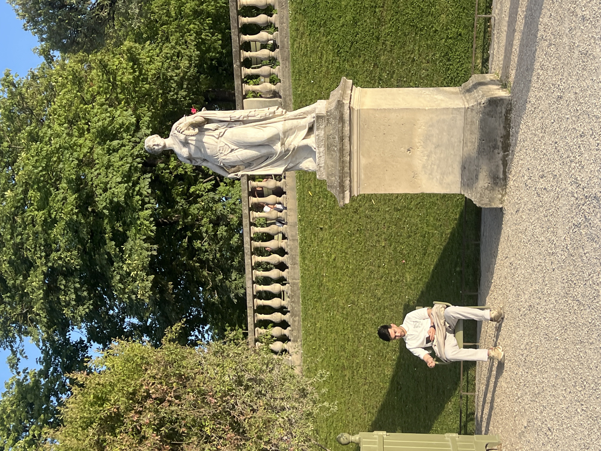 The image depicts a man sitting on a chair in a park, looking at his phone. He is wearing a white polo shirt and light-colored pants. A statue of a woman is visible in the background, along with a stone railing and a grassy slope. The scene appears to be outdoors in a well-maintained park with trees and a clear sky. The lighting suggests it is late afternoon. The presence of a statue and formal garden elements points to a location like the Luxembourg Gardens in Paris, France. The man is the primary human subject, engaged in a solitary activity. The statue serves as a prominent inanimate subject, a piece of public art within the park's landscape.