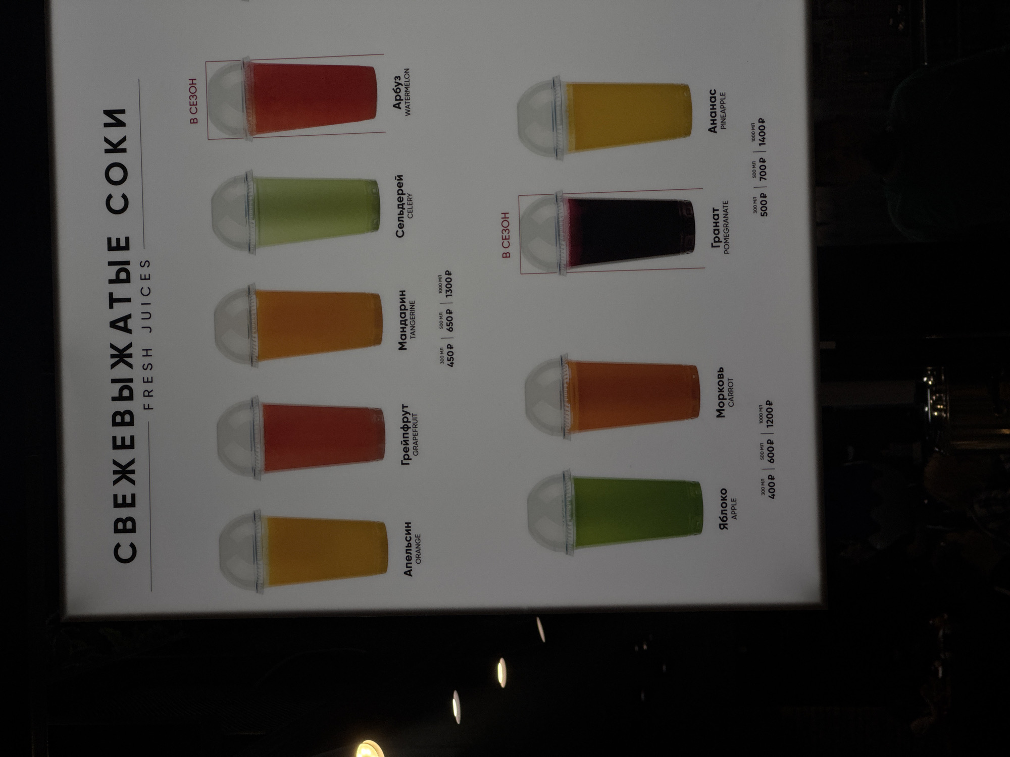 This image shows a sign advertising fresh juices, likely in a shop or cafe in Moscow, Russia. The sign displays various juice options with accompanying pictures, names in both Russian and English, and prices for different sizes (300ml, 500ml, and 1000ml where available). The juice options are: orange, grapefruit, apple, tangerine, celery, watermelon, carrot, pomegranate, and pineapple. "В СЕЗОН" means "In Season" is written next to the watermelon and pomegranate options. The text "СВЕЖЕВЫЖАТЫЕ СОКИ" translates to "Freshly Squeezed Juices".