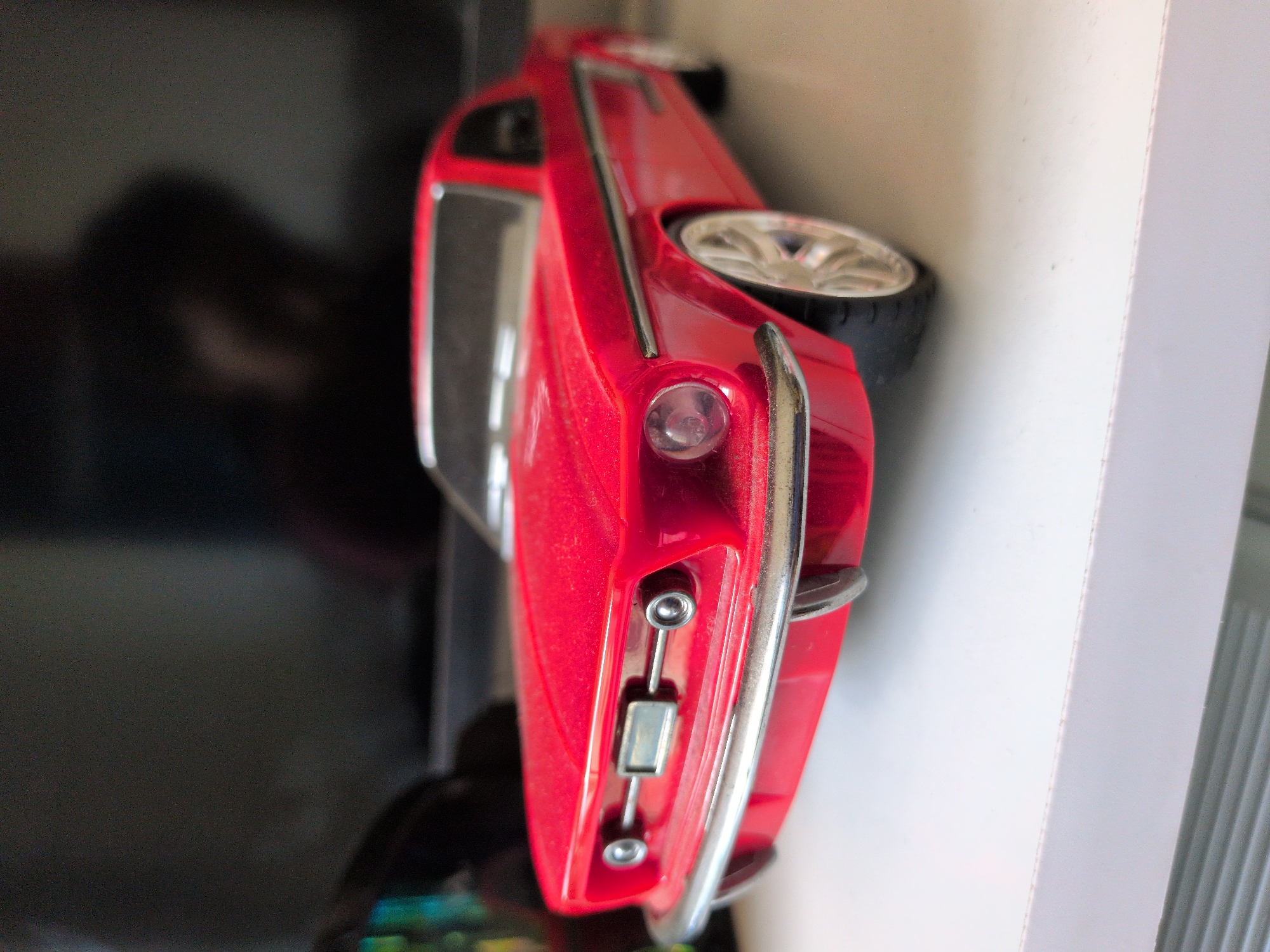A red toy car, resembling a classic American muscle car, sits on a white surface. The car is oriented at an angle, facing towards the left of the frame. Its front bumper and headlights are visible, as well as a portion of its side and a silver alloy wheel. The background is dark and out of focus, appearing to be a screen or a wall. There is no indication of the location being Mumbai, India.