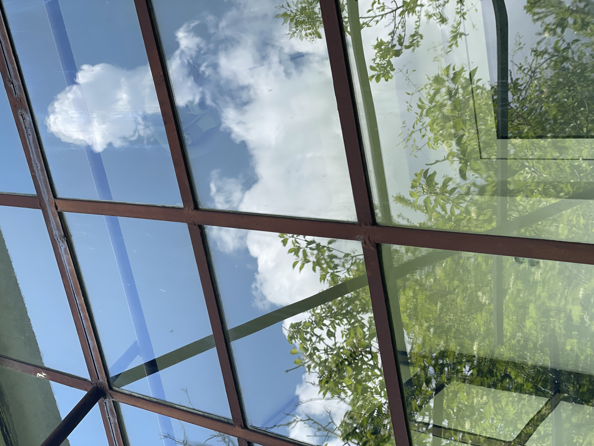 The image shows a close-up view of a windowpane with a grid of brown metal frames. The window reflects a bright blue sky with fluffy white clouds, as well as the green foliage of trees. The reflections are clear, suggesting a sunny day. The scene is photographed from a low angle, looking upwards through the window. The location is identified as Rudartsi, Bulgaria, though the image itself does not contain explicit geographical markers. It appears to be an exterior shot of a building, possibly a greenhouse or a structure with a large window surface. There are no people or animals visible in the image. The overall impression is one of nature and sky reflected on a man-made structure.