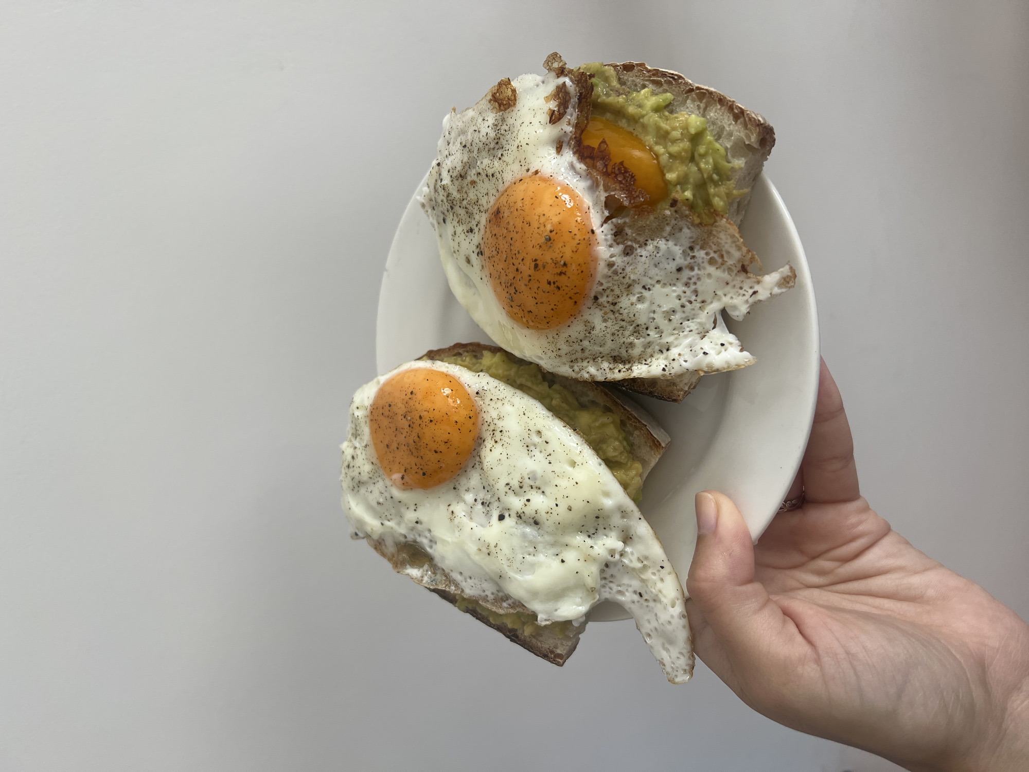A close-up, high-angle shot captures a person's hand holding a white plate featuring two servings of avocado toast with fried eggs. The background is a plain, light grey or white surface, suggesting a minimalist or studio-like setting.
Each serving on the plate consists of a slice of toasted bread, generously covered with mashed green avocado. Resting on the avocado are two perfectly fried eggs, distinguished by their vibrant orange, seemingly runny yolks and crisp white edges. Both eggs are visibly seasoned with a liberal amount of freshly ground black pepper. One egg, positioned towards the top right, has a slightly broken yolk, revealing a hint of the avocado beneath, while the other yolk, in the foreground, appears intact. The person's hand is visible on the right side of the frame, holding the edge of the round white plate.
The image portrays a simple yet appealing meal, likely breakfast or brunch. While the scene itself is minimalist, this meal is presumably prepared or enjoyed in the Nam Natural Heritage Area Trang, Vietnam, as per the provided context. There are no specific time-of-day indicators or weather cues visible in the image, nor is there any discernible text.