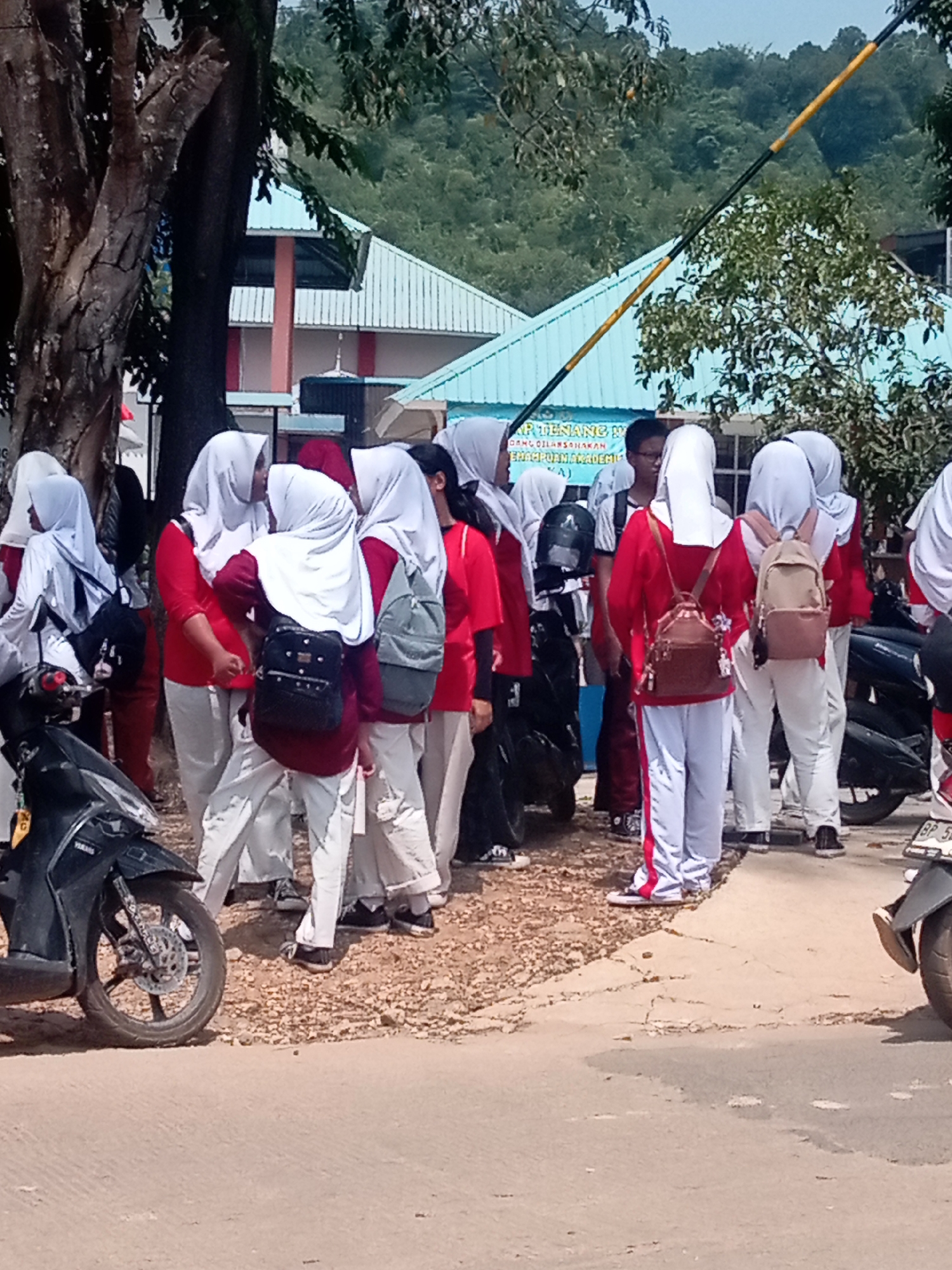 The image shows a group of young people, likely students, standing on a dusty path in front of a building with a light blue roof. Many of the individuals are wearing white hijabs and red shirts with white pants. Some are carrying backpacks in various colors. A black motorcycle is parked on the left side of the image. In the background, a lush green hill is visible under a bright sky. The scene appears to be a school campus. The building in the background has text which may be a sign for a school or educational institution. Based on the visual cues such as the architecture and the general environment, this location is likely in Batam, Indonesia.