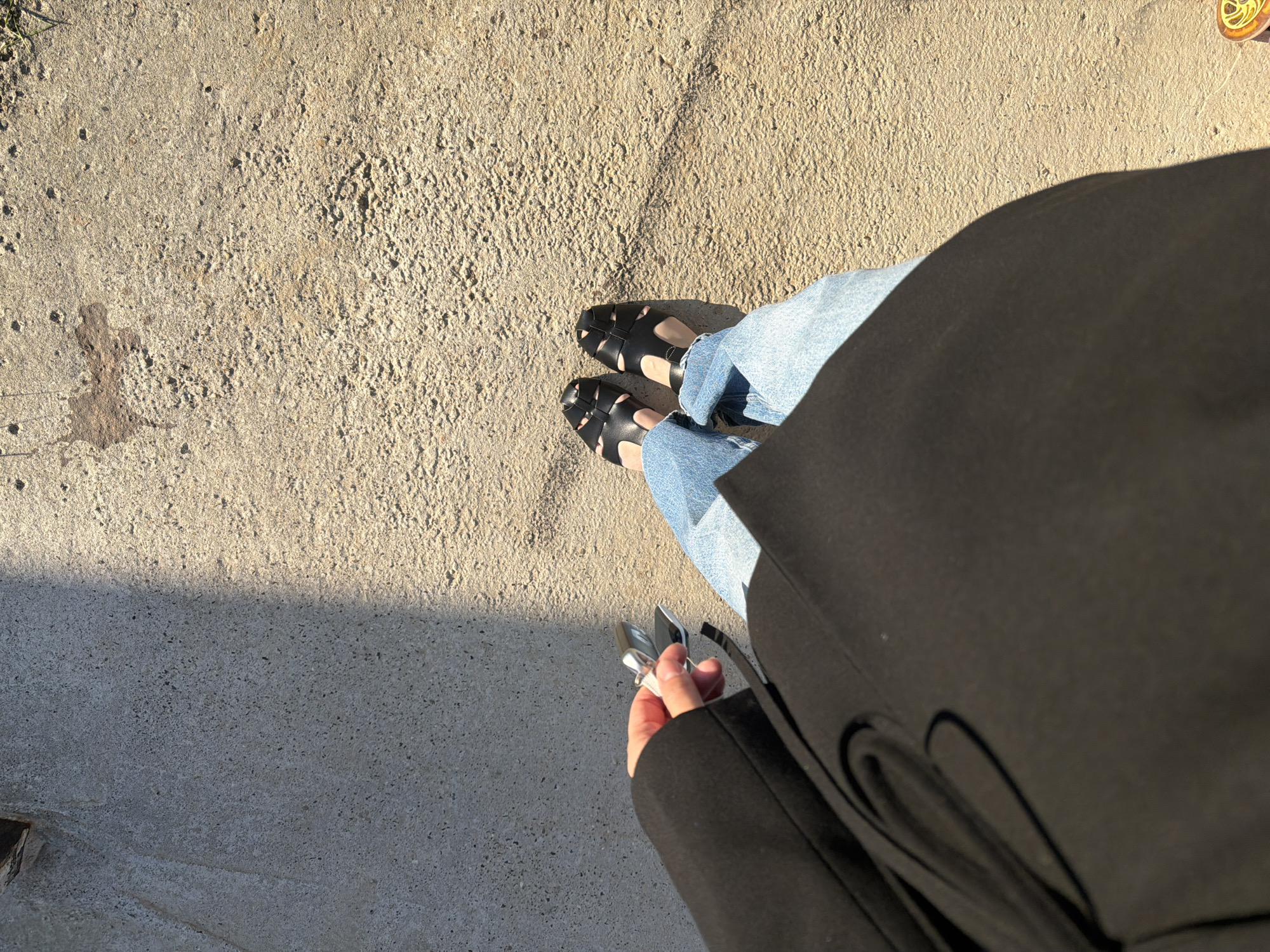 A first-person perspective shot looking down at a person standing on a concrete surface, possibly in the city of Chaikovsky, Russia, given the prompt. The person is wearing light-wash jeans and black sandals, with the feet positioned centrally. A black coat or jacket is partially visible in the bottom right, obscuring a portion of the lower body. The person's hand is holding keys and is positioned in the bottom left of the frame. The concrete ground occupies the majority of the image and shows a transition between a lighter, sunlit area in the upper right and a darker, shaded area in the lower left.
