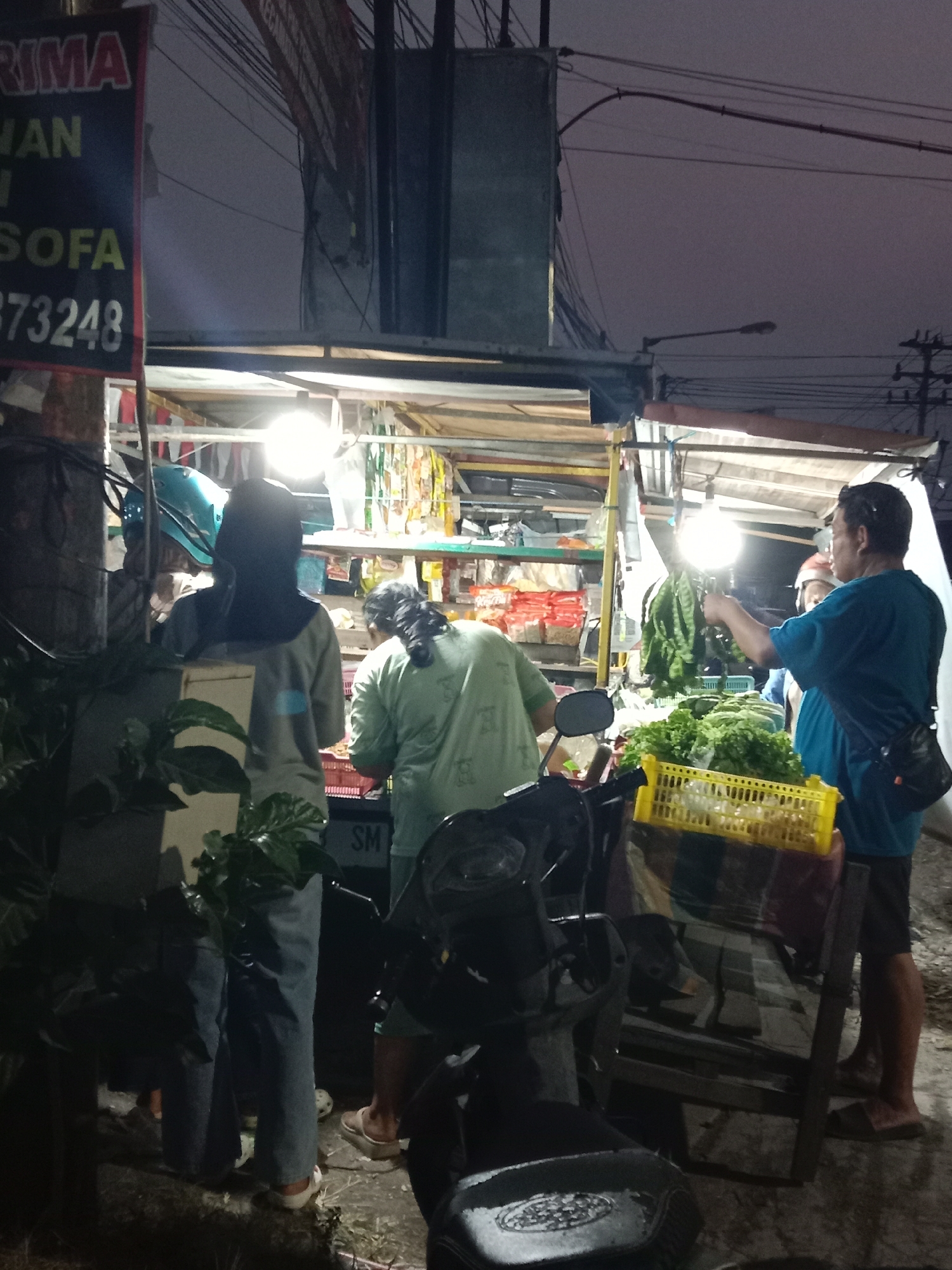 The image depicts a nighttime scene at a roadside stall in Langensari, Indonesia. A bright light illuminates the stall, revealing a variety of produce for sale, including leafy greens and what appear to be string beans or similar vegetables. Several individuals are present. One person, wearing a blue shirt and shorts, is standing and holding a bunch of vegetables. Another person, wearing a light green shirt, is partially visible and appears to be interacting with the produce. Two other individuals, wearing dark clothing and head coverings, are also present, their backs to the camera. A motorcycle is parked in the foreground. A sign with text, partially obscured by darkness and the angle, is visible on the left side of the image, with the words "RIMA," "WAN," "SOFA," and "373248" discernible. The environment suggests a bustling urban or semi-urban setting at night, likely a market or food stall area.