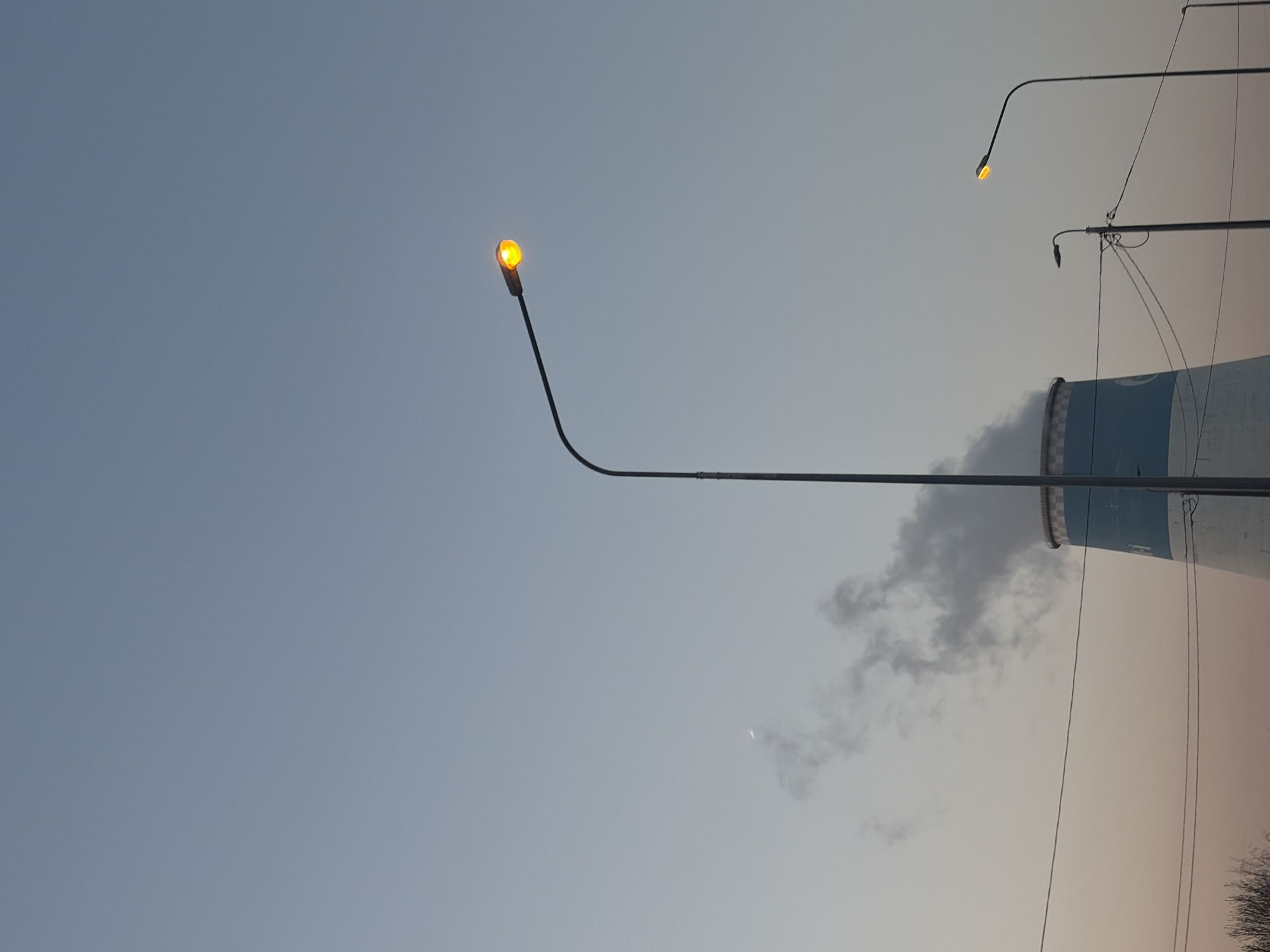 The image shows a cityscape at dusk in what is likely an industrial area. The sky is a pale blue-gray color. Two streetlights are visible, both with glowing orange bulbs. A cooling tower, characteristic of power plants, dominates the right side of the frame; it has a white and blue exterior. Some smoke or steam emanates from the top of the tower. There are power lines and the very top of a tree at the lower right. A sliver of the crescent moon is visible against the sky. Given the architectural style of the cooling tower and the general urban layout, and the presence of cooling towers on the horizon it is suggestive of Saint Petersburg, Russia, although it can not be definitively proven from the image.