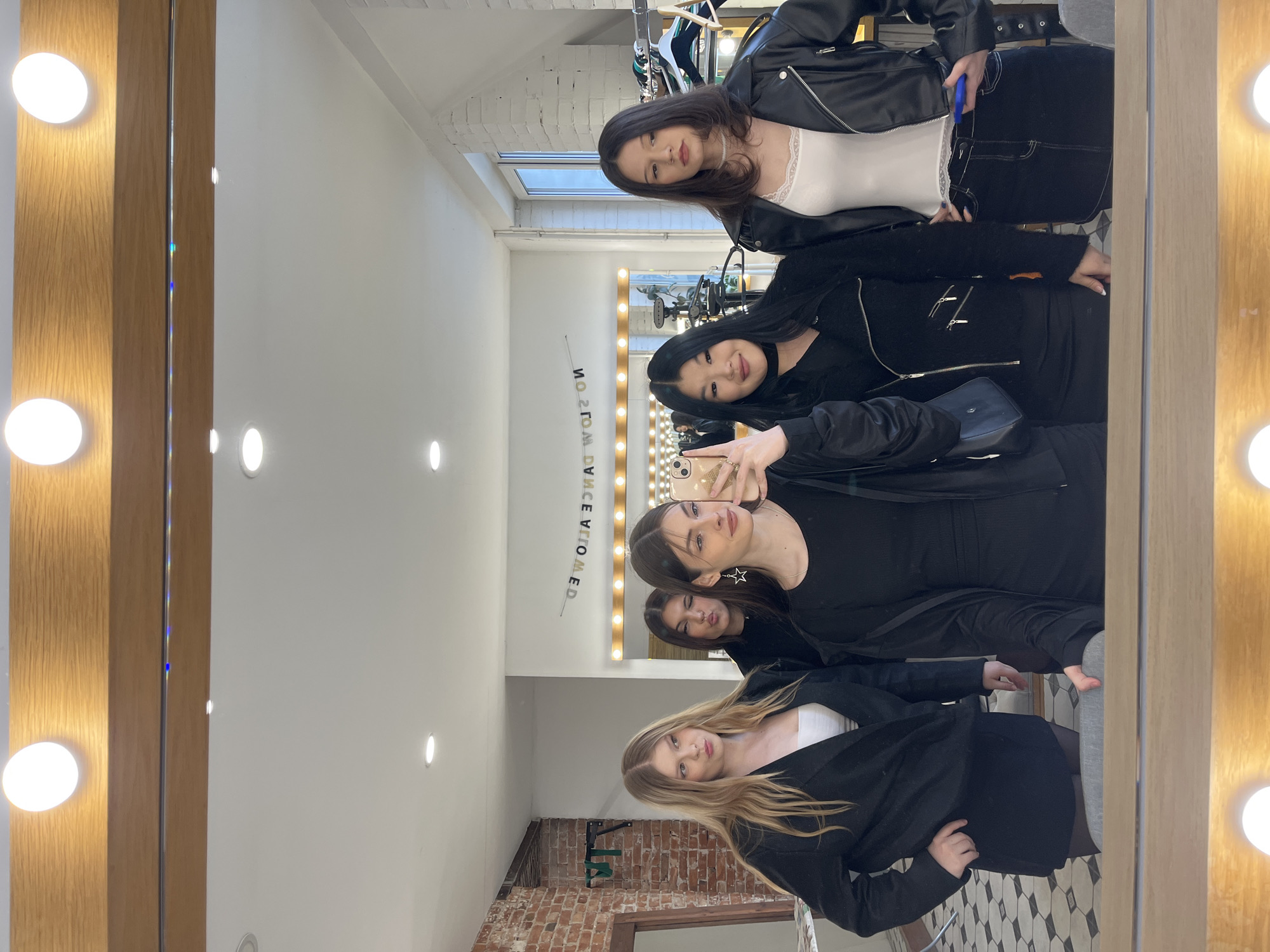 The image captures a mirror selfie of five young women inside what appears to be a studio or dressing room, located in Saint Petersburg, Russia. The photograph is rotated 90 degrees counter-clockwise from an upright perspective.
**Subjects:** Five young women, reflecting in a large mirror. The woman at the top of the current frame has dark hair and wears a white lace-trimmed top under a black leather jacket. Below her, another woman with dark hair holds a smartphone, capturing the reflection. A third woman, also with dark hair, makes a playful pouting face. Further down, another dark-haired woman is visible. The woman at the bottom of the current frame has long blonde hair and wears a black jacket over a white top.
**Objects:** A large vanity mirror with a light wooden frame and several integrated circular lights. A smartphone (likely an iPhone) is held for the selfie. Clothing racks with hangers are visible in the background reflection. A black shoulder bag is also seen. The interior features industrial-style ceiling lights, a section of exposed brick wall, and a patterned floor with hexagonal tiles.
**Activity or event taking place:** The women are posing for a group mirror selfie, seemingly during a daytime gathering or visit to a studio, salon, or fashion-related space.
**Notable details:**
- **Time of day:** Appears to be daytime, indicated by the natural light coming through a window visible in the background reflection.
- **Location cues:** The setting combines a modern, bright interior with industrial elements (exposed brick, ceiling fixtures). The vanity mirror suggests a space for makeup or dressing.
- **Emotions:** The women display relaxed, posed, or playful expressions.
- **Orientation:** The original image is rotated 90 degrees counter-clockwise, making the women appear horizontal within the frame.
**Visible text:** The reversed text "ЯЭƆИAD WOHƧ OИ" is visible on a sign or ledge in the background reflection, which reads "NO SHOW DANCER" when corrected.