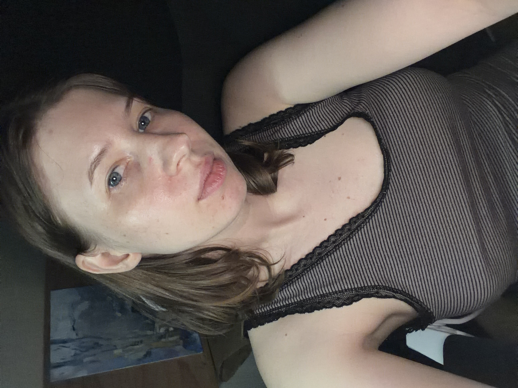 A young woman with light skin, blue eyes, and brown hair is looking at the camera. She is wearing a dark-colored, striped tank top with lace trim. A painting is visible in the background. The lighting is dim, suggesting an indoor setting, possibly at night. The exact location in Dongducheon-si, South Korea, is not discernible from the image.
