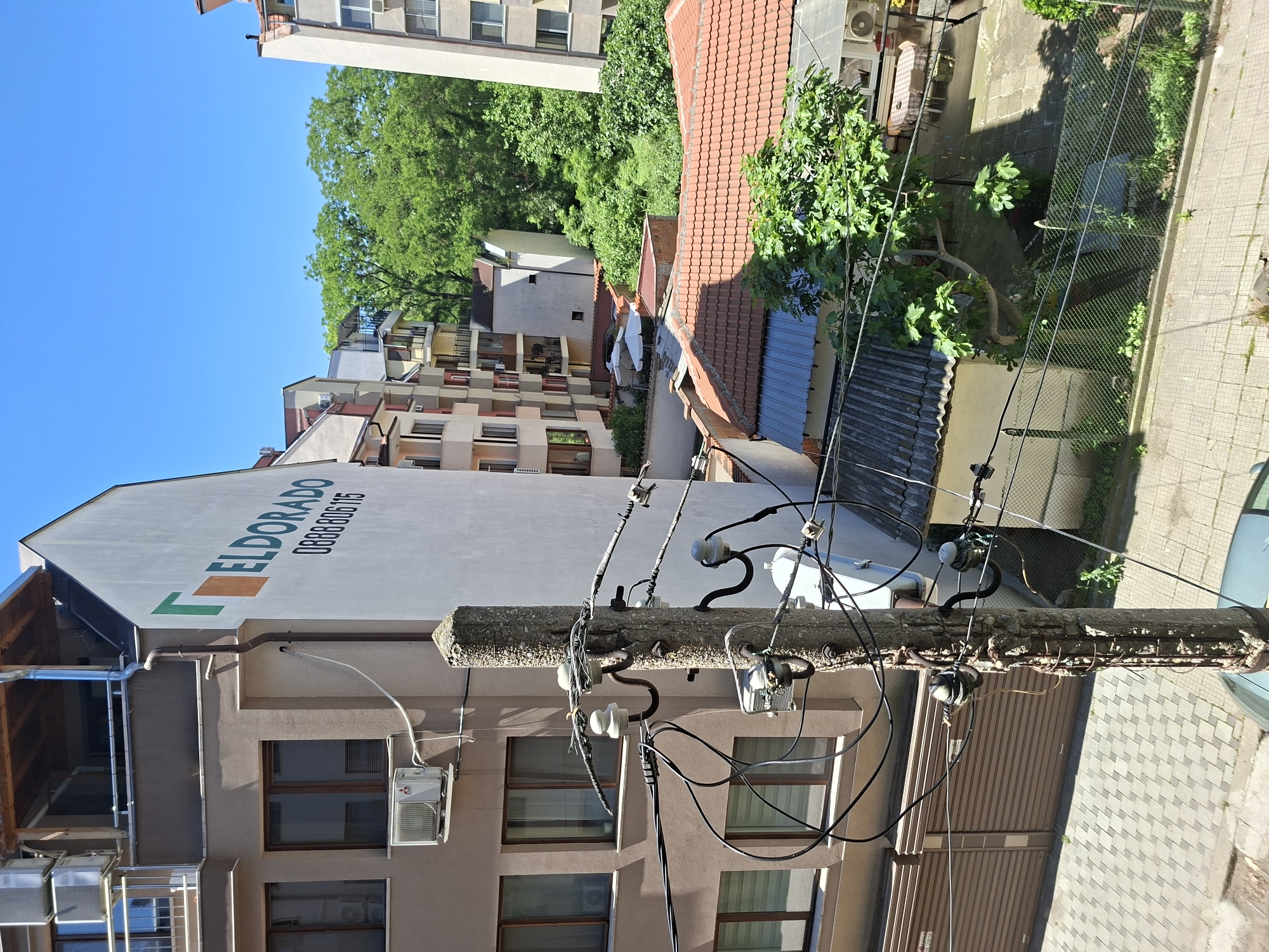 The image, taken outdoors, shows a view in Plovdiv, Bulgaria, predominantly featuring urban infrastructure. A weathered utility pole runs centrally through the frame, heavily burdened with electrical wires and insulators. Behind it is a building painted tan, with a sign on the roof reading "ELDORADO" and a phone number, along with windows and air conditioning units. The background reveals a diverse urban landscape, including buildings of various heights and architectural styles, some with red-tiled roofs. Green trees intersperse the buildings, providing a touch of natural contrast. To the right, a metal fence encloses a yard with vegetation. The image suggests an older part of the city with a mix of residential and commercial structures and established utility infrastructure. The lighting is bright, indicating a clear, sunny day. No people are visible.