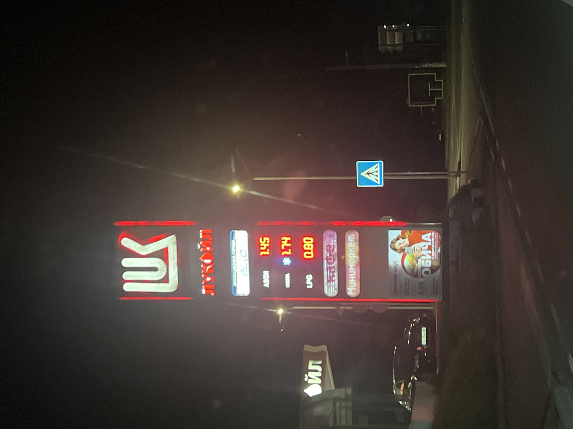 The image depicts a Lukoil gas station in Banichan, Bulgaria at night. The station's illuminated sign displays the prices for different fuels: A95H at 1.45, Diesel at 1.74, and LPG at 0.80. Other signage, including a promotional advertisement, is also visible. A dark-colored vehicle is parked in front of the station. A road sign is near the station.
