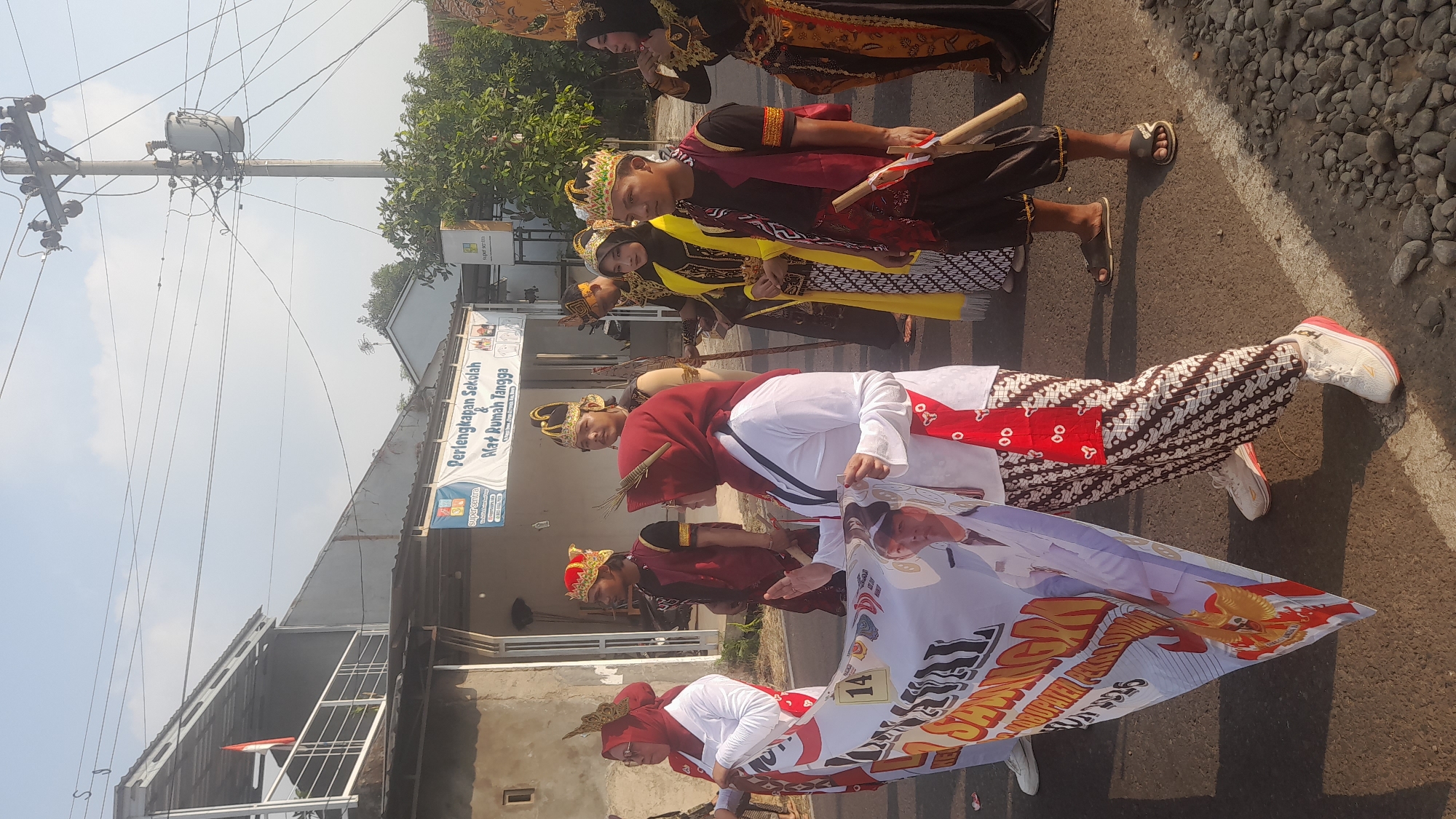 In Lemahabang, Indonesia, a procession is underway on a paved road with a rocky shoulder, likely during daylight given the shadows. Several individuals, dressed in traditional Indonesian attire, are participating. On the left, two women in white shirts and red hijabs are holding a large banner that features text and an image of a man. One woman's lower body is visible, wearing patterned batik trousers and white sneakers. To their right, a group of young men are also in traditional dress, some wearing elaborate headdresses and vests over shirts. One man in the foreground wears a yellow and black outfit with a headdress and holds wooden sticks. Behind them, a building with a corrugated metal roof and a sign reading "Perlengkapan Sekolah & Alat Rumah Tangga" is visible, indicating a school and home goods store. Further back, a utility pole with a transformer stands against a clear sky. The scene suggests a cultural event or parade.
