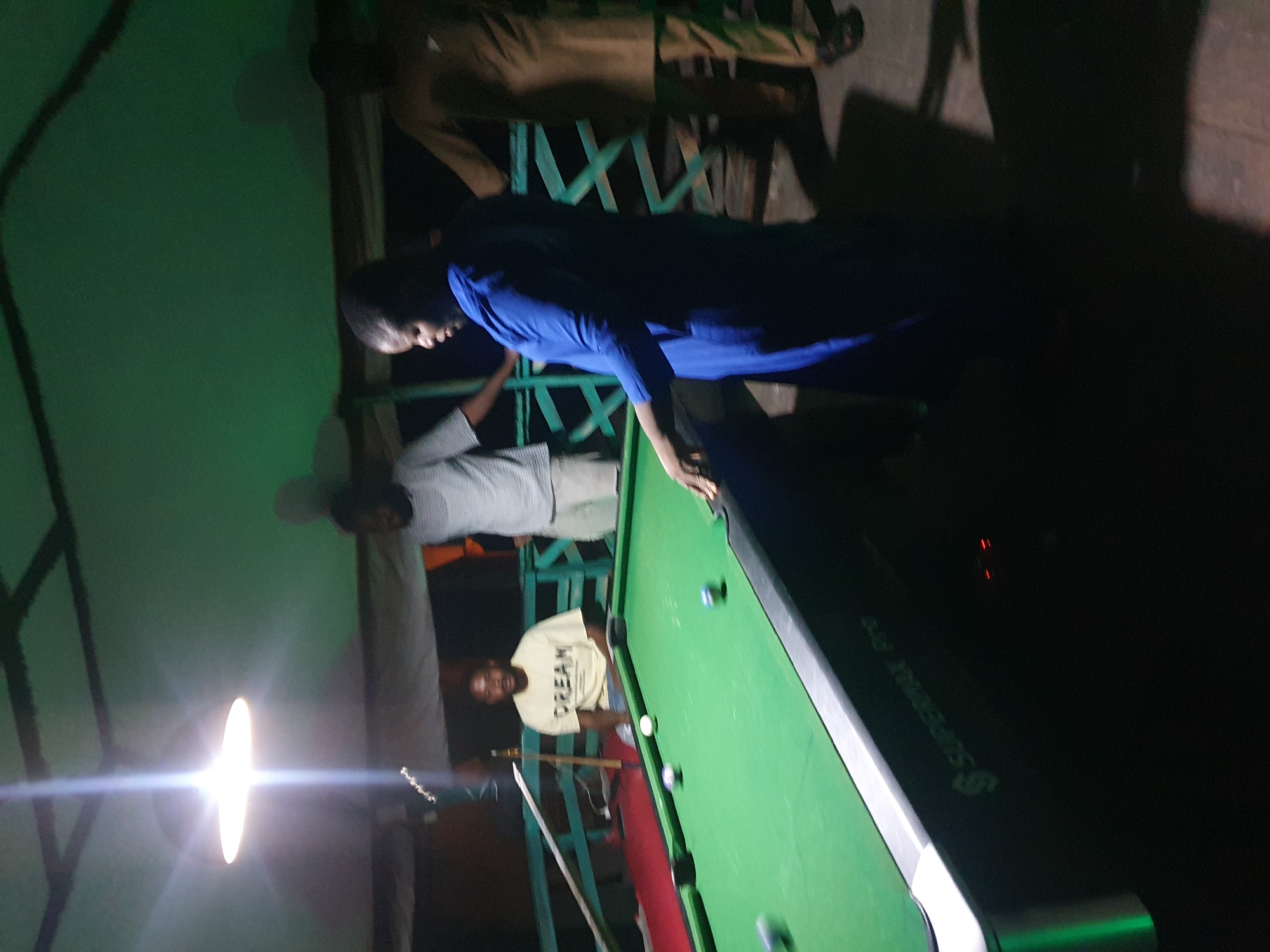 The image shows a group of people playing billiards at night. The scene appears to be an outdoor or semi-outdoor setting, possibly a bar or a recreational area, given the presence of a pool table and a green wall. Three men are visible, two of whom are actively engaged with the pool table. One man, wearing a blue shirt, is leaning over the table, seemingly preparing to take a shot. Another man in a light-colored shirt with the word "DREAM" printed on it is watching the game. The third man, dressed in a gray and white striped shirt, is standing further back, observing the players. A bright light source, likely a lamp, illuminates the area, casting strong shadows and creating lens flare. The overall atmosphere seems casual and social. Some of the pool balls are visible on the green felt of the table. The word "DREAM" is clearly legible on the t-shirt of one of the men. The time of day is night, indicated by the artificial lighting. The location context is Lokoja, Nigeria.