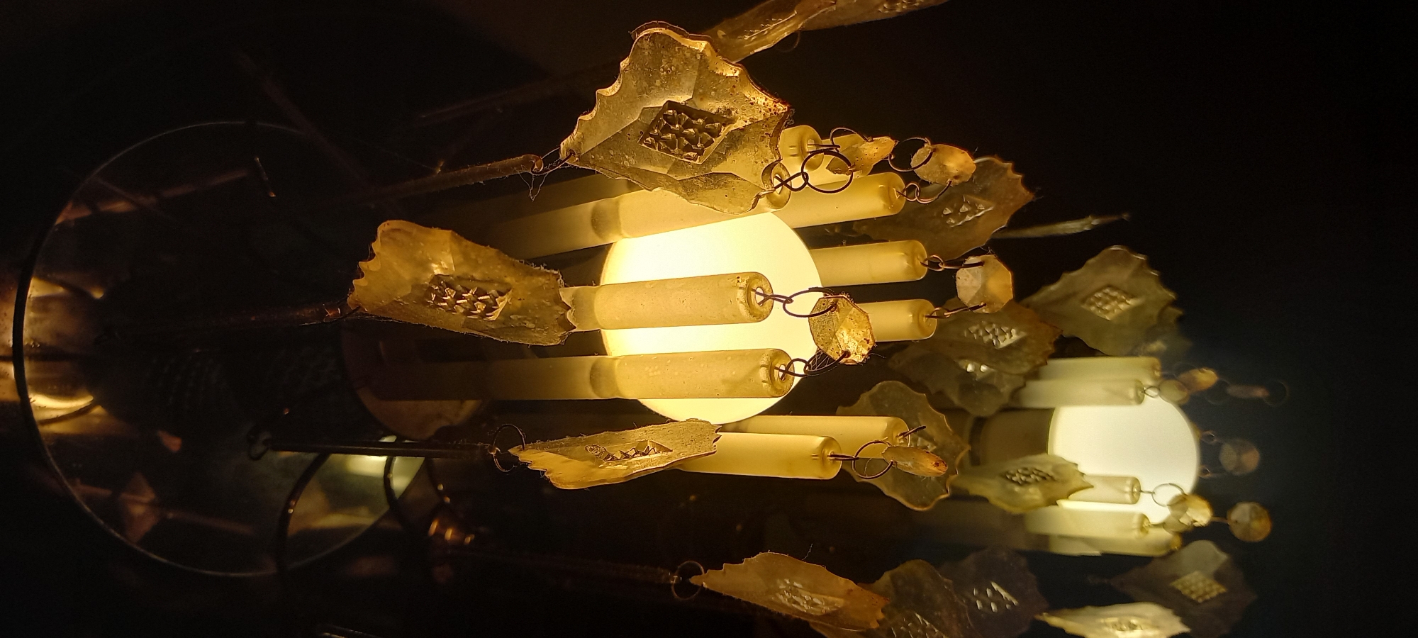 An upward-angled photograph depicts an illuminated decorative light fixture, likely a chandelier or pendant lamp, against a dark background. The central element is a bright, spherical light source emitting a warm, yellowish-white light. Surrounding this light are multiple translucent, cylindrical elements, vertically suspended, appearing to be made of frosted glass or plastic. Densely hung below these cylinders and around the fixture are numerous flat, irregularly shaped decorative pieces, resembling stylized leaves or abstract forms. These pieces are translucent, possessing a yellowish-brown hue, and many feature embossed or etched diamond-like patterns centrally, with some exhibiting scalloped edges. They are attached by small metal rings or wires. Interspersed among these flat pieces are smaller, irregular, rough-cut, clear or semi-clear, stone-like elements, also suspended by metal rings. The upper portion of the fixture, above the main light, appears metallic and reflective, showing subtle reflections of the light source and structural components. The immediate environment is dark, indicating an indoor setting with the fixture as the primary light source. There are no individuals, discernible actions, or interactions present in the image. The image contains no visual cues or identifiable landmarks to determine its specific location within Kryvyi Rih, Ukraine.