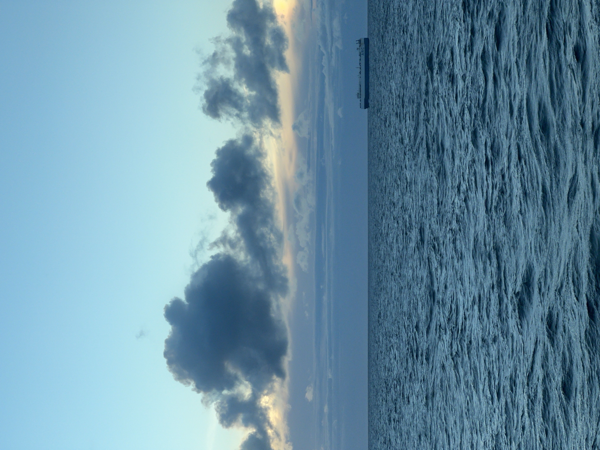A cargo ship, identified by the "ARC" logo on its side, sails on a calm, dark blue sea under a pale blue sky. The sky is filled with a mix of dark, heavy clouds and lighter, wispy formations, with a hint of yellow light peeking through the clouds on the right horizon. The sea exhibits gentle ripples and waves, reflecting the muted colors of the sky. The image is a static, horizontal composition with the horizon line dividing the frame roughly in the middle. There are no people visible in the frame. The location is unknown.