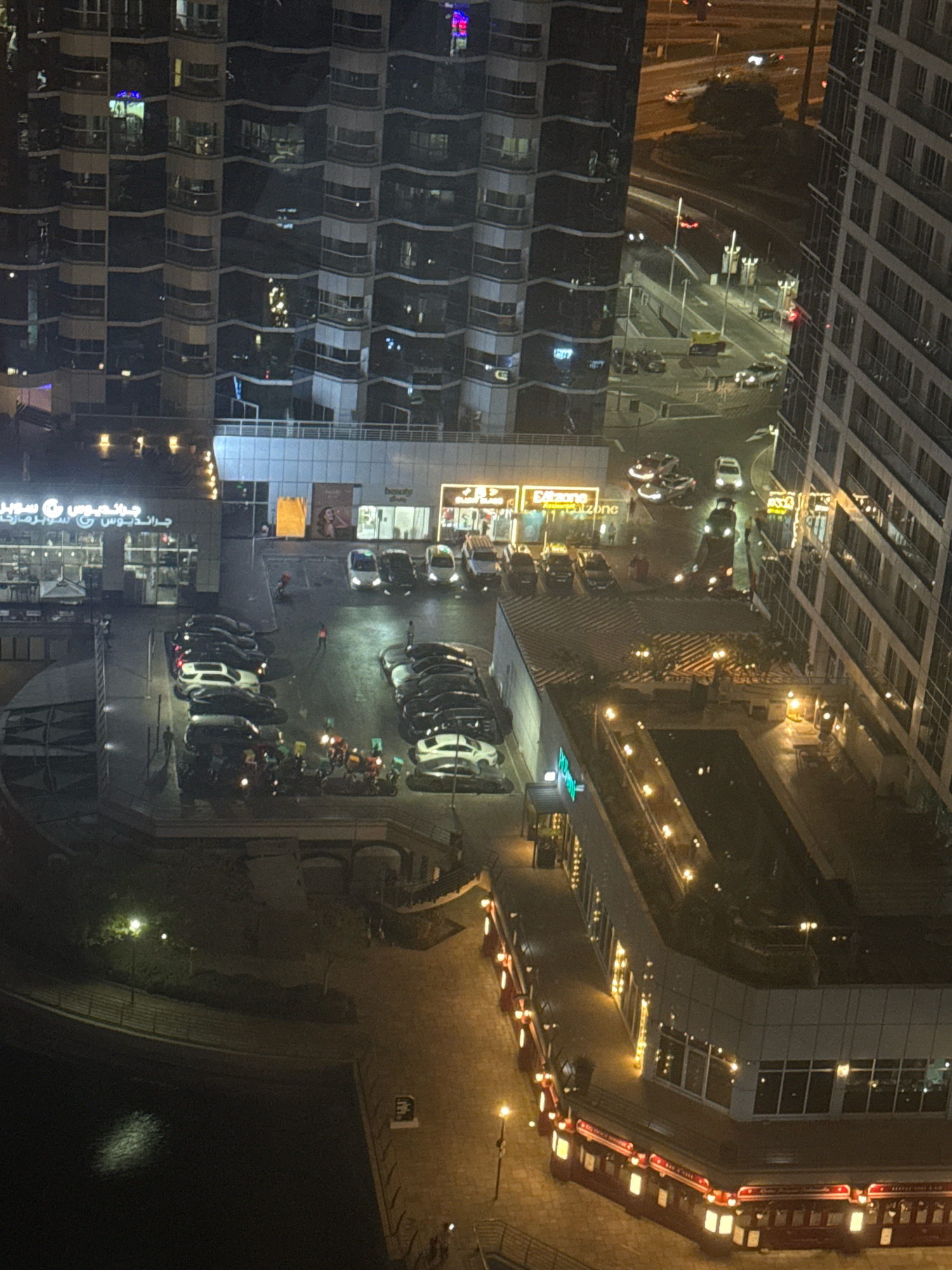 The image captures a nighttime view of a waterfront area in Dubai, United Arab Emirates, likely a commercial or residential district with high-rise buildings and a canal or marina. In the foreground, a multi-story building with illuminated windows and balconies is visible. Below it, a plaza area features several parked cars and a group of motorcycles, some with riders, possibly engaged in a gathering or delivery service.
Further into the scene, a street with cars in motion and parked vehicles is observed, bordered by commercial establishments with glowing signs, including what appears to be a supermarket and retail stores. The overall environment is characterized by artificial lighting from buildings, streetlights, and vehicle headlights, reflecting on the wet pavement, suggesting recent rain or the presence of water. The architecture suggests a modern urban setting.