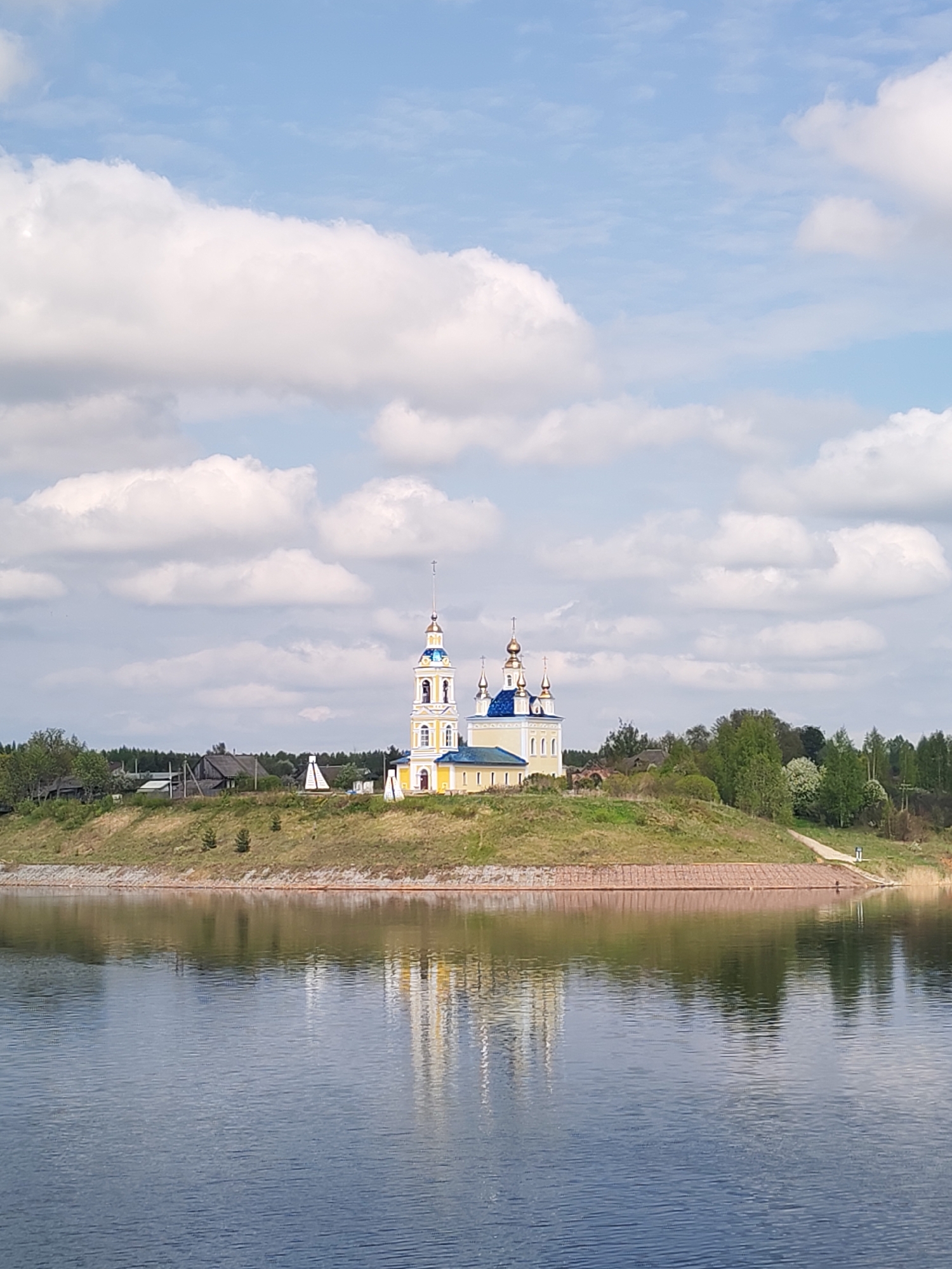 The image shows a yellow and blue church with golden domes standing on a grassy hill, reflected in the calm water below. The church is likely an Orthodox church, given the architectural style. The surrounding area includes trees and houses on the hilltop behind the church. The sky is blue with fluffy white clouds. The location is Глебово, Russia.