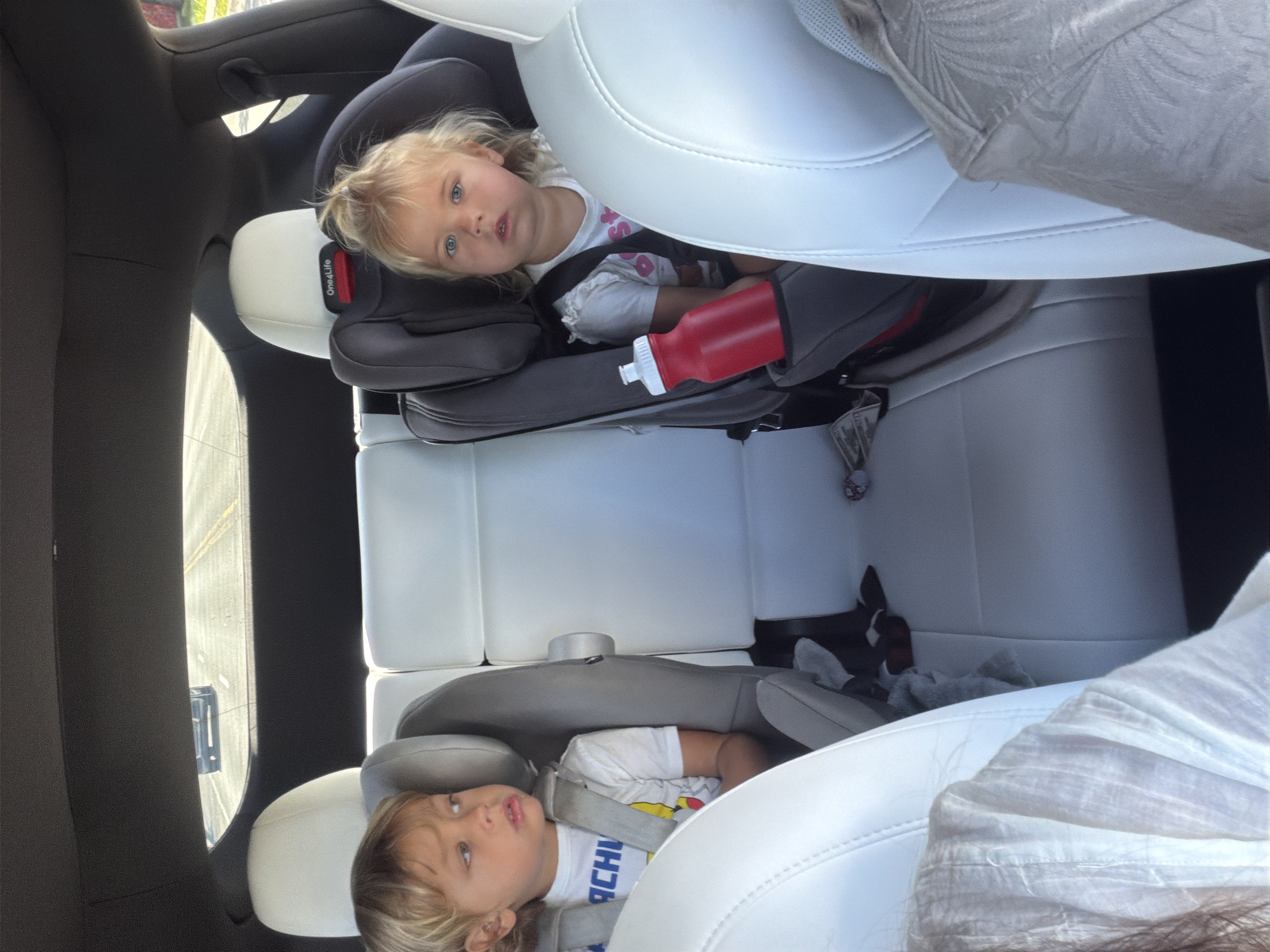 The image shows the interior of a car, likely a Tesla, with two young children in car seats in the back. The child on the left, a boy with blonde hair, is wearing a white t-shirt with blue lettering and is looking to his left. The child on the right, a girl with blonde hair and blue eyes, is wearing a white t-shirt and is looking forward. She has a red water bottle in her car seat. The car seats are grey and are positioned on white leather seats. Through the rear window, a highway is visible with other cars. The scene suggests a family road trip or everyday travel. The location is not explicitly stated to be Miami, Florida, but the bright sunlight and the style of the car interior are consistent with a warm climate.