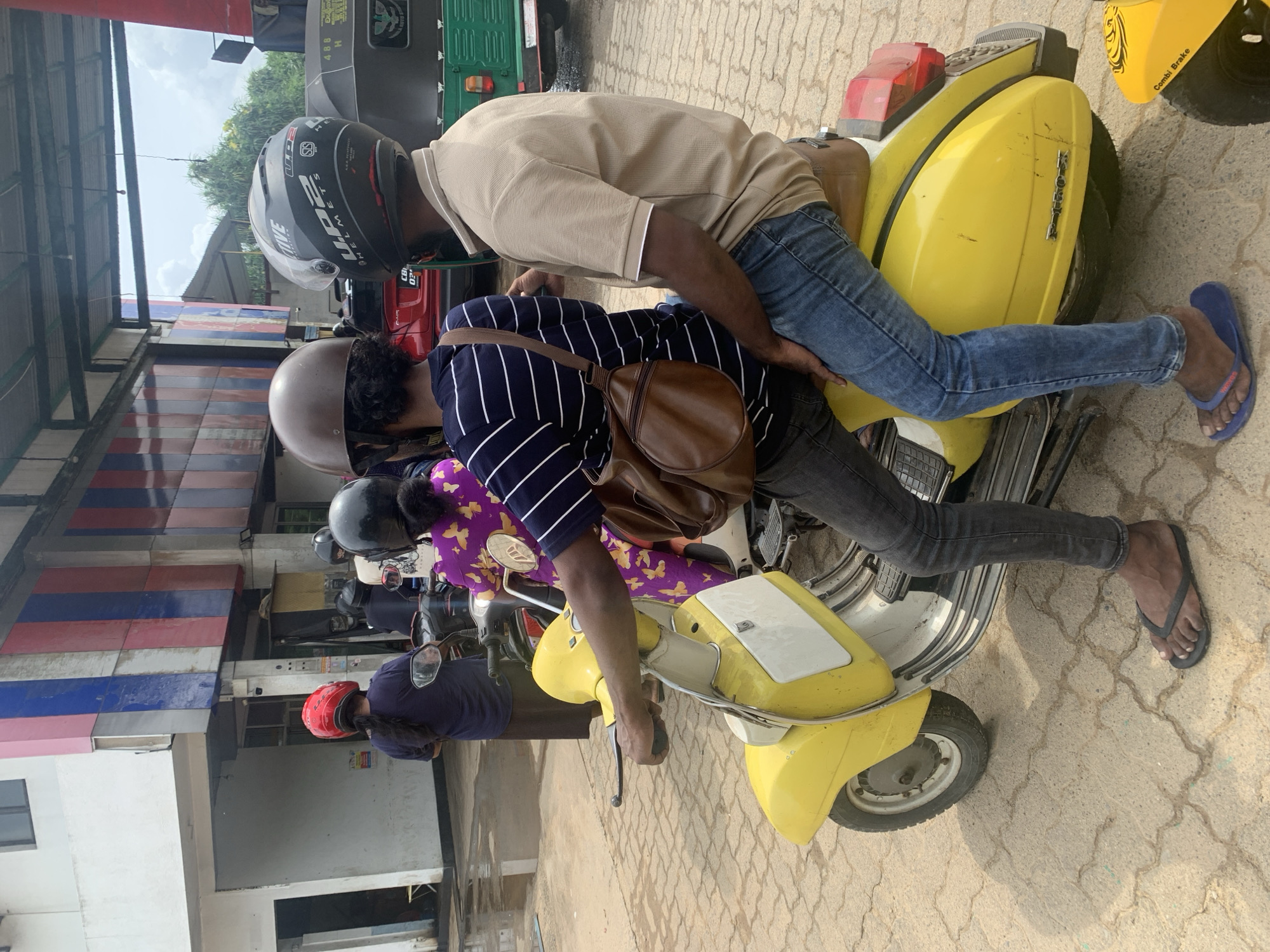In Goviyapana, Sri Lanka, a scene captures two people on yellow scooters parked on a paved area. One scooter is occupied by a person wearing a tan shirt and blue jeans, as well as a helmet. The second scooter has a person with a striped shirt, a brown bag, jeans, and a helmet. Further back in the image is a person with a purple shirt and another with a red helmet, each on scooters. In the background, there are buildings with blue, red, and white facades, as well as other vehicles.