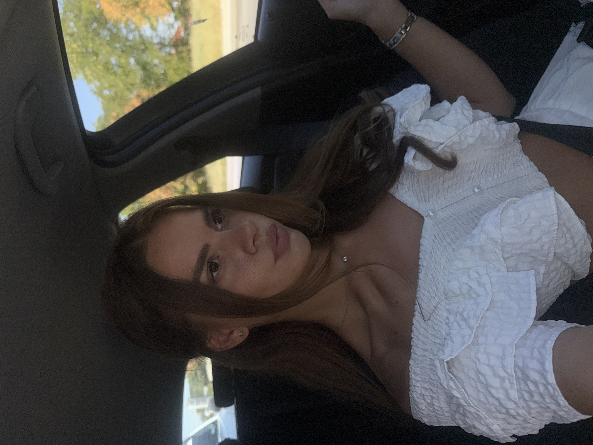 A young woman with brown hair is seated in the passenger seat of a car. She is wearing an off-the-shoulder white ruffled top and a silver necklace with a small pendant. Her right arm rests on the armrest, and she is wearing a silver bracelet. The background visible through the car window shows trees with autumn foliage, suggesting the image was taken in the city of Plovdiv, Bulgaria, during the fall season. The interior of the car is dark, with the ceiling and seats visible. The lighting is natural, coming from the window.