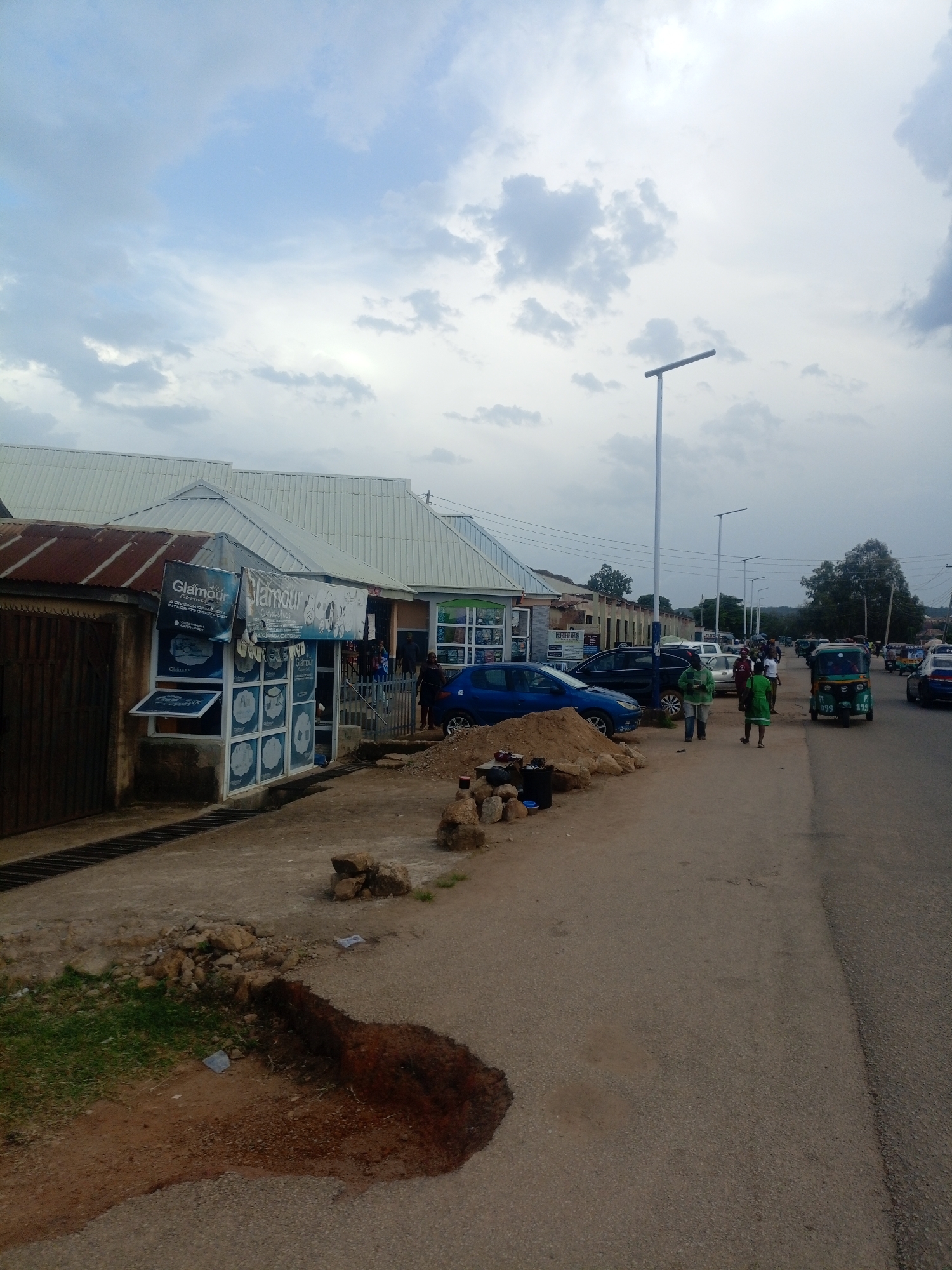 The image captures a street scene in Jos, Nigeria, under an overcast sky. The foreground features a dirt patch with scattered rocks and a small area of exposed brown earth, suggesting a construction or disrepair zone. To the left, a building with a corrugated iron roof and a wooden gate stands adjacent to a shop with a modern facade displaying "Glamour Cosmetics & Beauty". Several windows of this shop are open, revealing shelves stocked with products. A blue car is parked in front of the shop.
Moving towards the right, the scene opens up to a paved road where several vehicles are present, including a tricycle (Keke Napep) in the mid-ground and other cars further down the road. A group of people, some dressed in casual clothing, are seen walking along the side of the road. The buildings lining the street appear to be a mix of commercial establishments with some residential structures in the background. Streetlights are visible along the road, indicating urban infrastructure. The overall impression is that of a busy commercial area in a Nigerian city.