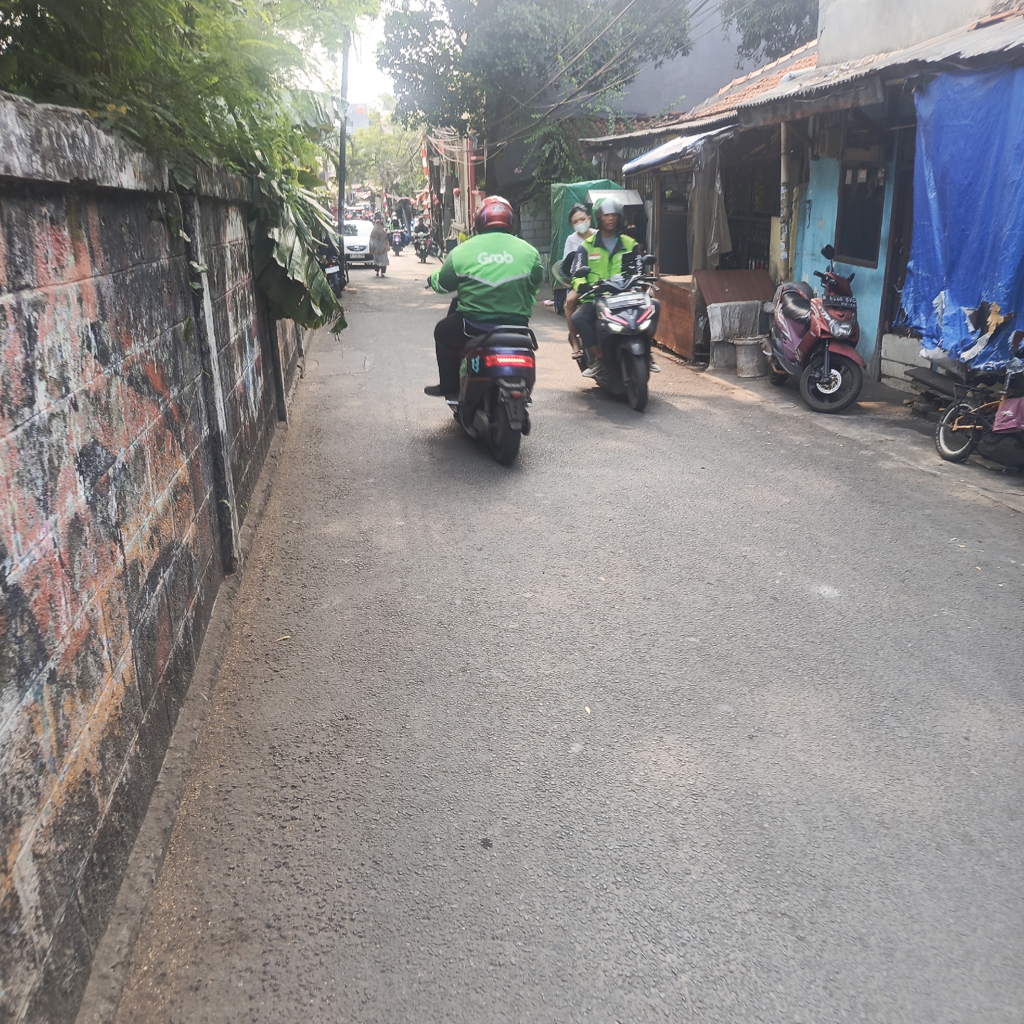 An outdoor, eye-level, medium shot captures a narrow, paved alleyway in South Jakarta, Indonesia, on a bright, sunny day. The alley is lined on the left by a tall, weathered brick wall with graffiti. On the right, a row of small, rustic buildings with corrugated metal roofs and blue tarpaulin awnings stand adjacent to the alley. Several motorcycles are parked along the right side, and a few people are visible.
In the foreground, a person on a dark blue electric motorcycle, wearing a green jacket with "Grab" written on the back and a red helmet, is riding away from the camera. To the right of this rider, another motorcycle is occupied by two people, both wearing green jackets and helmets. The rider wears a light green jacket and a helmet with a white face mask. The passenger, a woman, wears a white face mask and a green shirt.
Further down the alley, more motorcycles and a white car are visible, along with other pedestrians. The environment is characterized by its urban, somewhat impoverished setting, with visible power lines overhead and lush green foliage peeking over the brick wall on the left. The overall impression is that of a typical busy street scene in a residential or commercial area of Jakarta.
