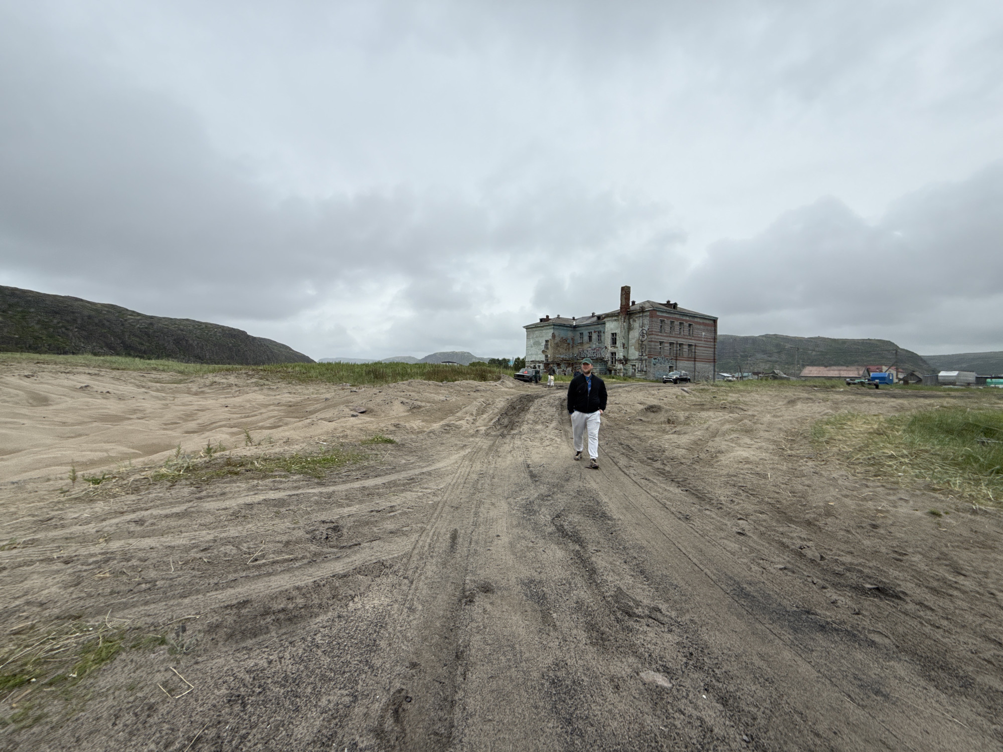 A man walks down a dirt road in Teriberka, Russia. The road is rutted and sandy, with some sparse vegetation growing along the sides. In the background, there is a large, dilapidated building, possibly an old dormitory or administrative complex, with broken windows and graffiti. The sky is overcast and gray, typical of the Arctic climate. Several vehicles are parked near the building. The landscape is barren and hilly, with a sense of desolation.