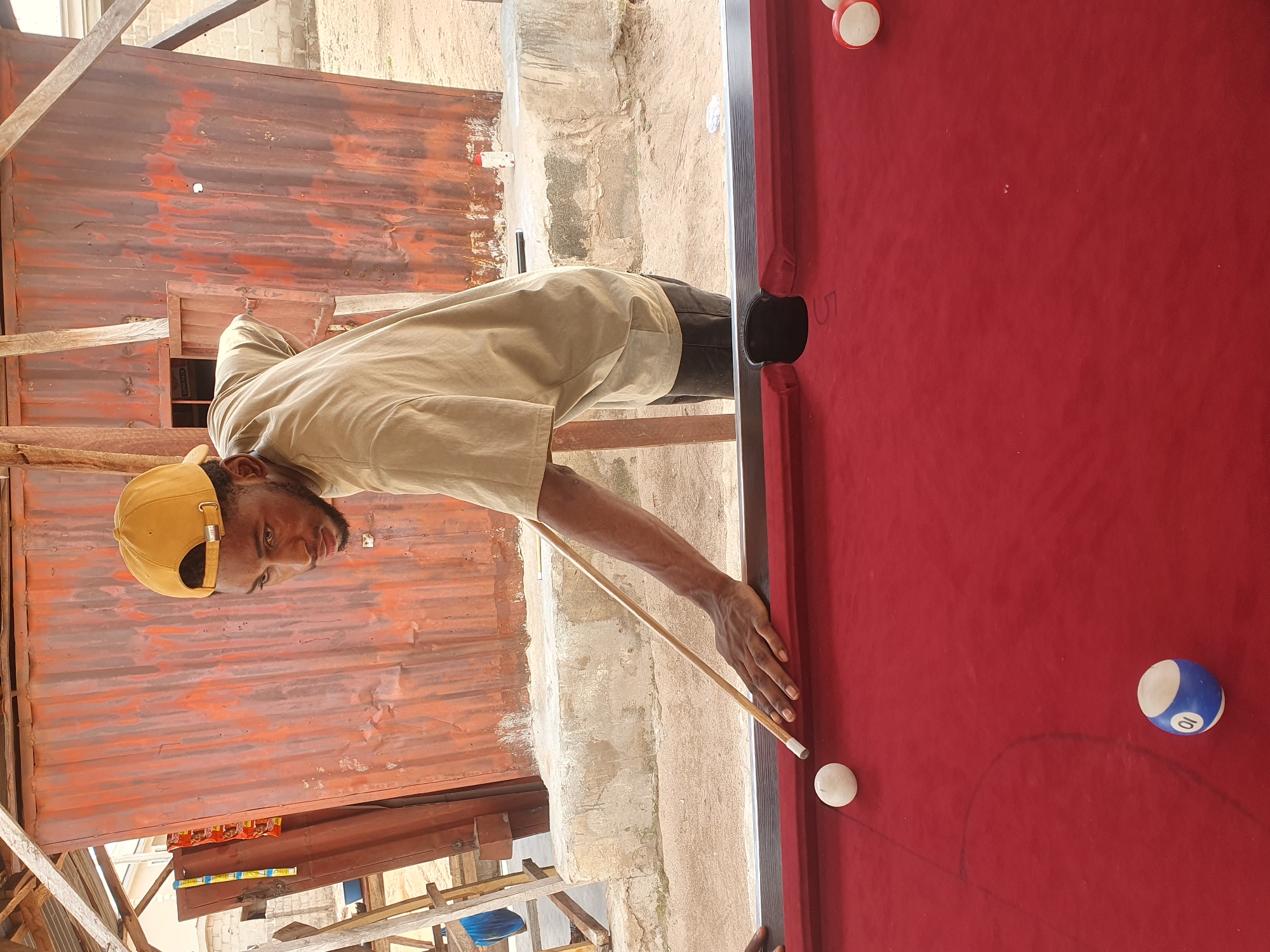 A young Black man wearing a tan t-shirt and a yellow baseball cap is playing pool in an outdoor setting that appears to be Lokoja, Nigeria. He is focused on striking a white cue ball with a pool cue, aiming for another ball on the red felt surface of the pool table. The background features a weathered, corrugated metal structure with a wooden frame, suggesting it's a makeshift or outdoor recreational area. The ground is sandy, with some concrete blocks visible, and sparse vegetation. One of the billiard balls on the table has the number '10' in a blue circle.