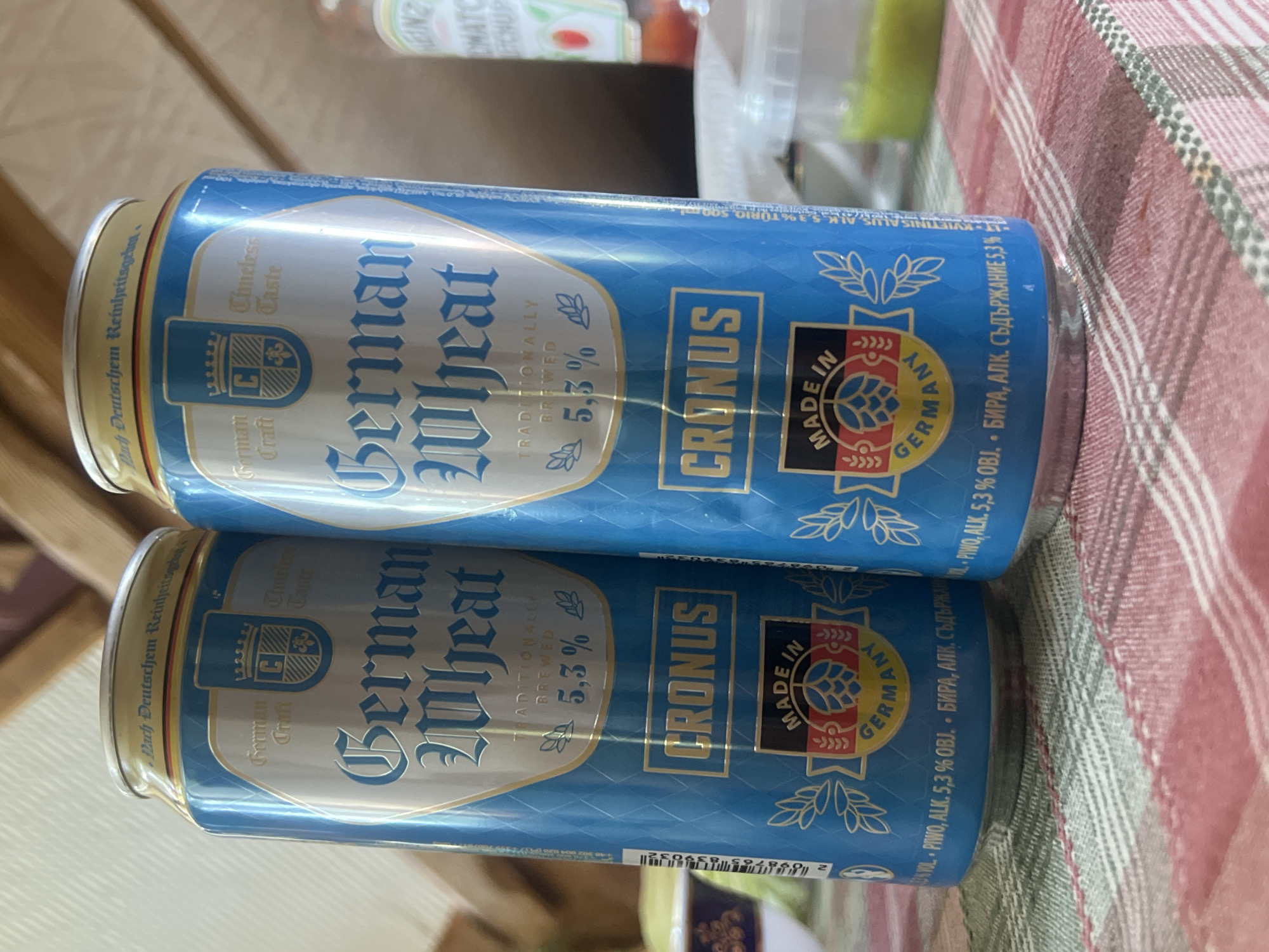 Two blue beer cans with yellow and silver labels are displayed on a checkered tablecloth. The cans are positioned side-by-side, with the left can slightly overlapping the right. Both cans have the text "German Wheat" in large, ornate font. Below this, "TRADITIONALLY BREWED 5,3%" is visible. A circular logo with the words "MADE IN GERMANY" and a hop illustration is present towards the bottom of each can, beneath the word "CRONUS". The background is blurred, but suggests an indoor setting with wooden elements and a hint of a bottle of ketchup in the far right. The specific location of Norviliškės, Lithuania is not discernible from the image itself.