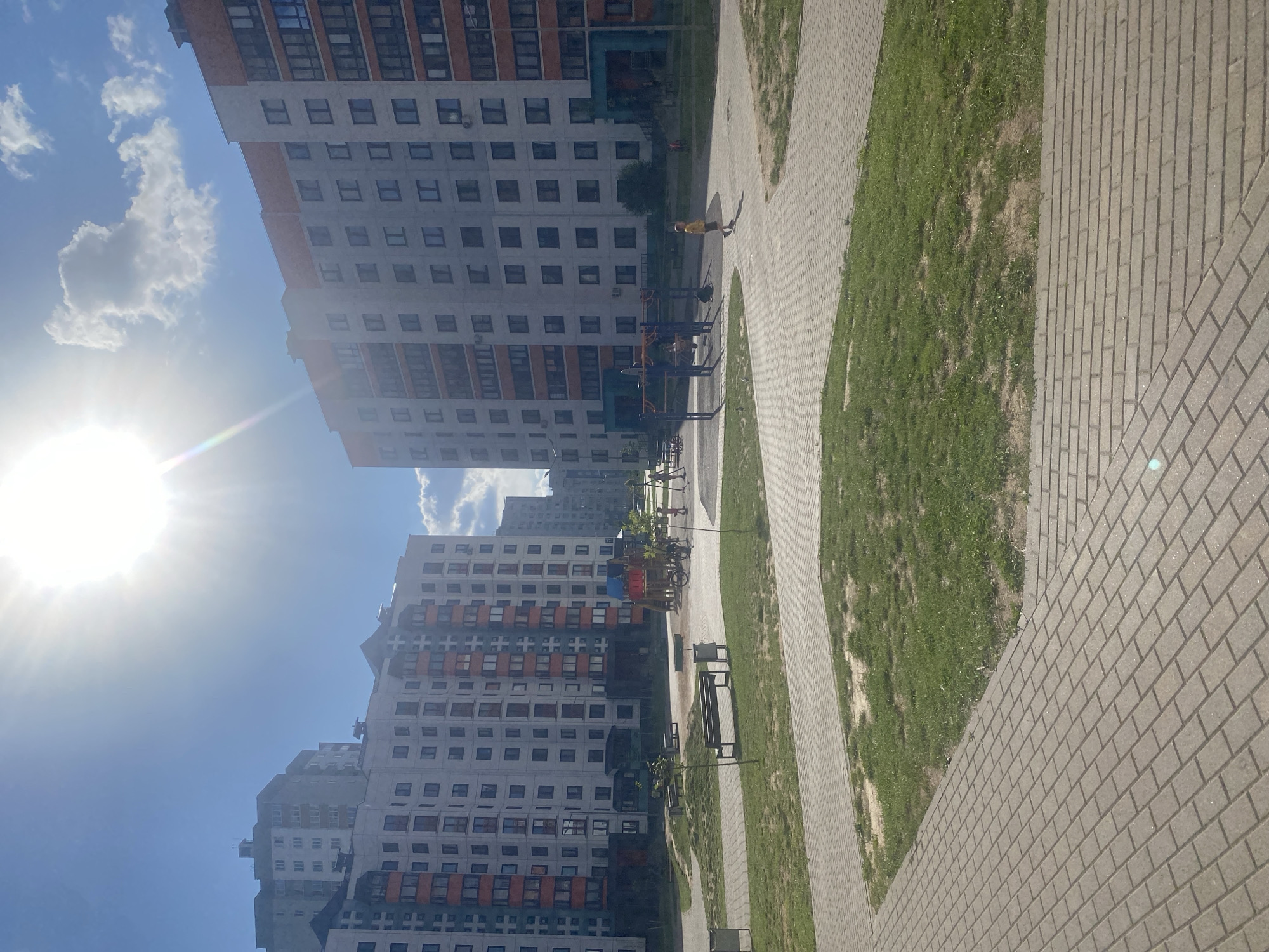 This image depicts a residential courtyard on a sunny day, likely in Minsk, Belarus, as suggested by its architectural style. In the foreground, a paved walkway made of grey brick leads the viewer into the scene. Beyond the walkway, a patch of green grass is visible, interspersed with some bare soil. Further back, more paved areas and grassy patches continue, creating a multi-level effect.
Dominating the midground and background are several large, multi-story apartment buildings. These buildings are modern in design, with light-colored facades and numerous windows, many of which appear to have enclosed balconies with orange accents. The buildings are arranged in a way that creates a courtyard space.
Within this courtyard, there are elements of a playground and recreational area. A small children's playground with colorful structures is visible, along with some exercise equipment in blue metal. A few children are present; one child in a yellow shirt is walking across the paved area, and other figures are visible near the playground. Benches are placed in various spots, suggesting a place for residents to relax.
The sky is a vibrant blue with a few white clouds. The sun is very bright, positioned high in the sky and causing a sun flare that obscures part of the image. The overall impression is of a relatively new, well-maintained urban residential environment.