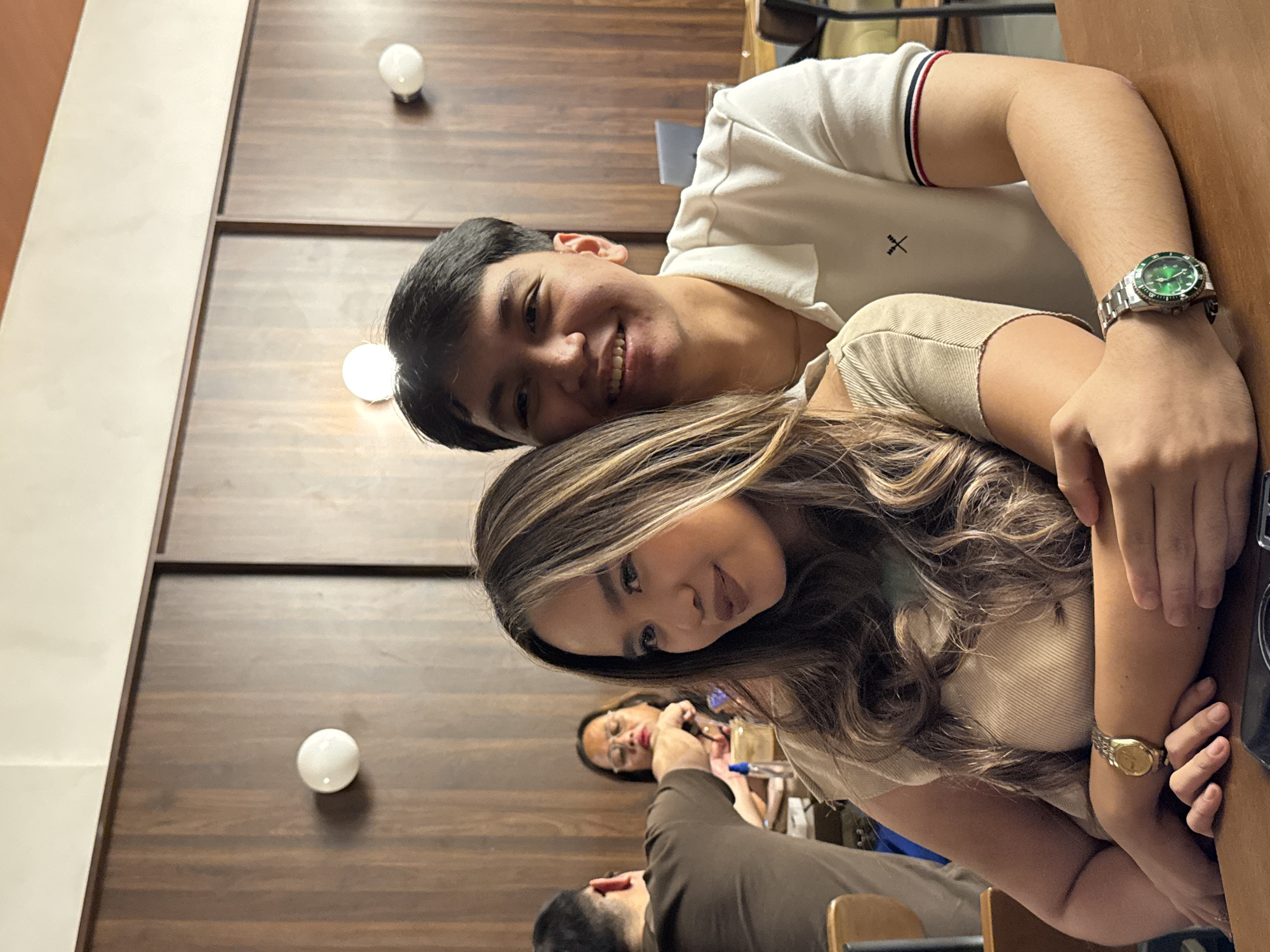 The image shows a young man and woman posing closely together, with the man's arm around the woman. He is wearing a white polo shirt with red and blue trim on the collar and sleeves, and a silver watch with a green face. The woman is wearing a tan-colored shirt and a gold watch. Her hair is long and wavy with blonde highlights.
In the background, there is a wooden wall with three round light fixtures. Two other people can be seen sitting at a table in the background. The image was taken indoors in Davao City, Philippines.