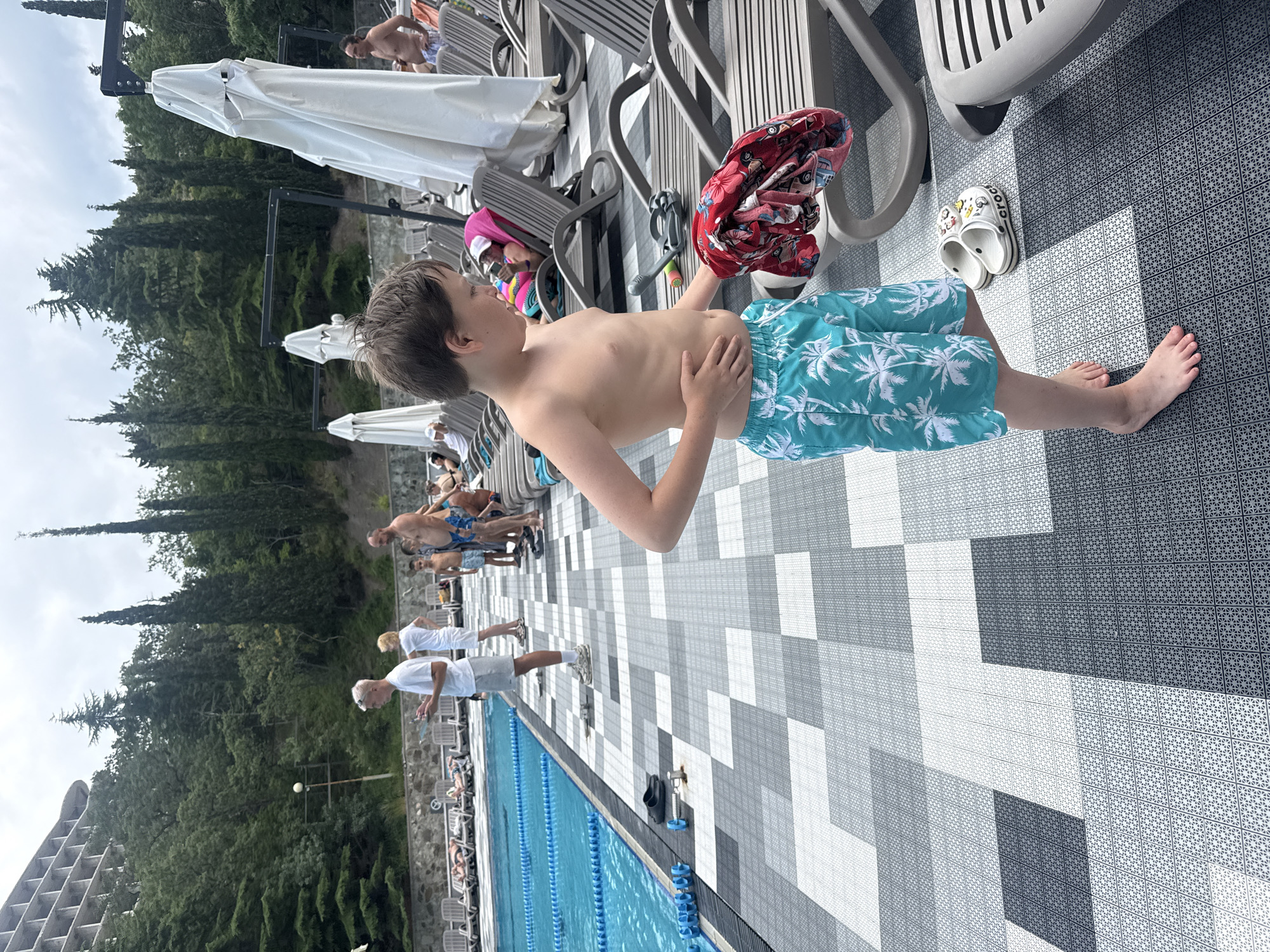 In a bright, outdoor setting with a swimming pool, a young boy stands on a patterned tiled deck. He is shirtless, wearing blue swim trunks with a white palm tree pattern. In his right hand, he holds a red and white patterned garment. To his right, on the tiles, are a pair of white Crocs-style sandals. The background shows more people in swim attire, lounge chairs, umbrellas, and lush green trees. A swimming pool with blue lane markers is visible to the left. The scene appears to be during daytime, likely a warm and sunny day, suggesting a vacation or recreational activity at a resort or hotel.