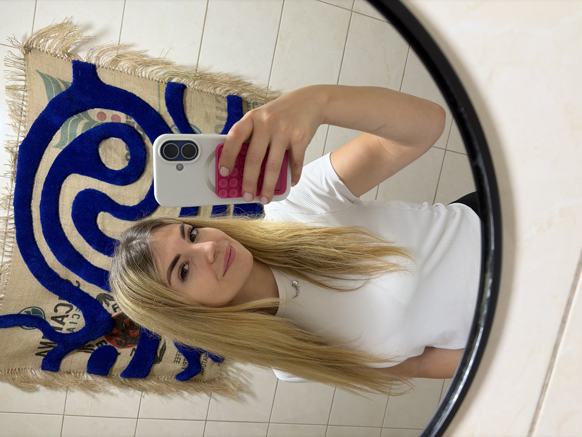 A young woman is captured taking a mirror selfie, seen from the chest up, in what appears to be an indoor setting with tiled walls and floor. She has long, straight blonde hair, fair skin, and is looking directly at the camera with a slight, pleasant smile. She is wearing a white, ribbed short-sleeved t-shirt and a small, delicate pendant necklace.
She holds a white smartphone with a distinctive pink pop socket or grip on its back. Her nails are painted a light pink. The selfie is taken in a round mirror, whose dark frame is visible on the right side of the image.
Behind the woman, on the tiled floor, lies a unique rug or mat. This rug features a prominent, textured blue swirling or organic design laid over a beige, burlap-like material. Visible text on the rug, which appears mirrored due to the reflection, clearly spells out "CAFE". Other fragmented, mirrored letters and words are discernible, suggesting it might be made from repurposed coffee sacks, with words like "MATURE" and "COFFEE" partially visible. The rug has frayed, fringed edges. The overall impression is casual and self-aware. The location context provided is Budva, Montenegro.