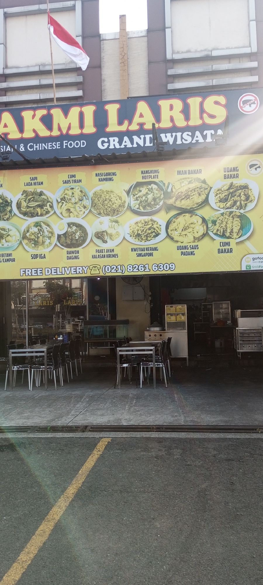 The image depicts the exterior of a restaurant named "BAKMI LARIS GRAND WISATA" located in Lambangjaya, Indonesia. The restaurant's signage is prominently displayed, featuring the restaurant name in large yellow letters on a dark background. Below the name, text indicates "CHINESE FOOD". A colorful menu with images of various dishes is visible, showcasing items like "SAPI LADA HITAM," "CUMI SAUS TIRAM," "NASI GORENG," "IKAN BAKAR," and more. The restaurant offers "FREE DELIVERY" and displays a phone number, "(021) 8261 6309." There is also a "GoFood" and "GrabFood" logo visible, suggesting delivery partnerships. The restaurant appears to have an outdoor seating area with several tables and chairs. The building facade is made of a material resembling concrete or stone. An Indonesian flag is flying from a pole above the restaurant. The overall scene suggests a commercial establishment in an urban or semi-urban setting in Indonesia.