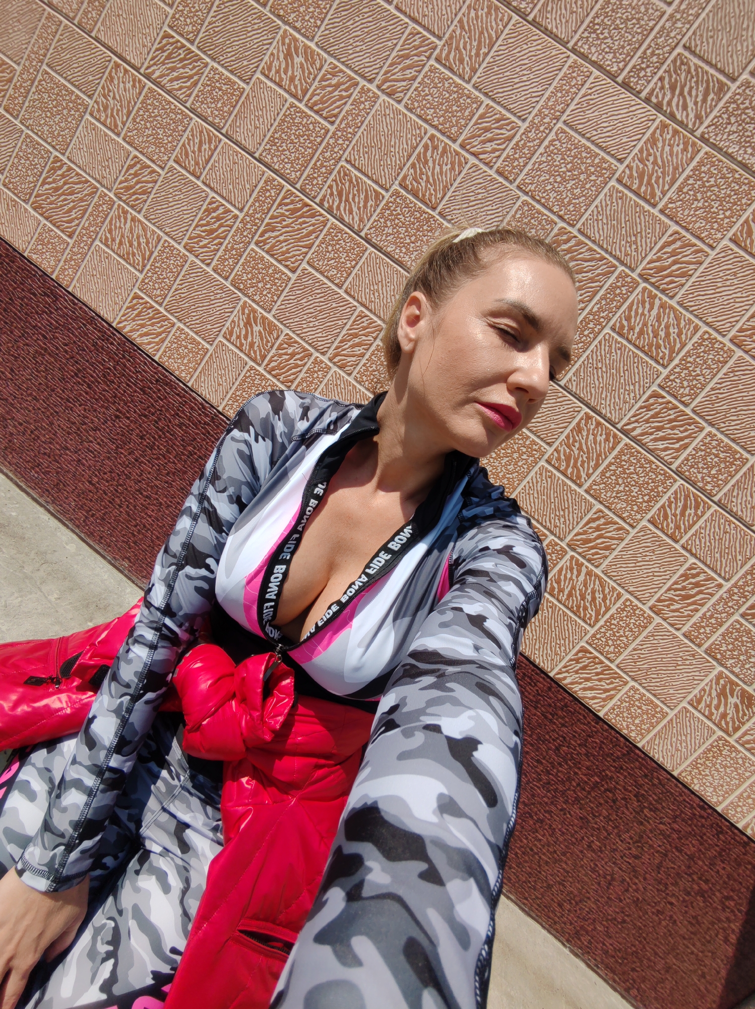 The image captures a female individual, likely an adult, taking a selfie outdoors in what is specified as Petropavlovsk-Kamchatsky, Russia, although no specific geographic landmarks are discernible to confirm this location. The woman's head is tilted, and her eyes are squinted or closed, possibly due to bright sunlight. Her hair, appearing blonde, is pulled back. She wears red lipstick. Her attire consists of a long-sleeved garment or suit featuring a grey, white, and black camouflage pattern. The top section of this garment has a deep V-neckline with a white base and a prominent pink stripe bearing mirrored text, which reads "BORN TO RIDE" (partially visible). A distinct bright red, shiny, and possibly padded garment is tied or wrapped around her waist. The background comprises an upper section of a diagonally patterned tiled wall in shades of light brown and beige, featuring alternating textured squares. Below this tiled wall, separated by a diagonal line, is a dark reddish-brown textured surface. A light grey ground surface is visible in the lower left portion of the frame. The scene is under strong direct sunlight, casting defined highlights and shadows.